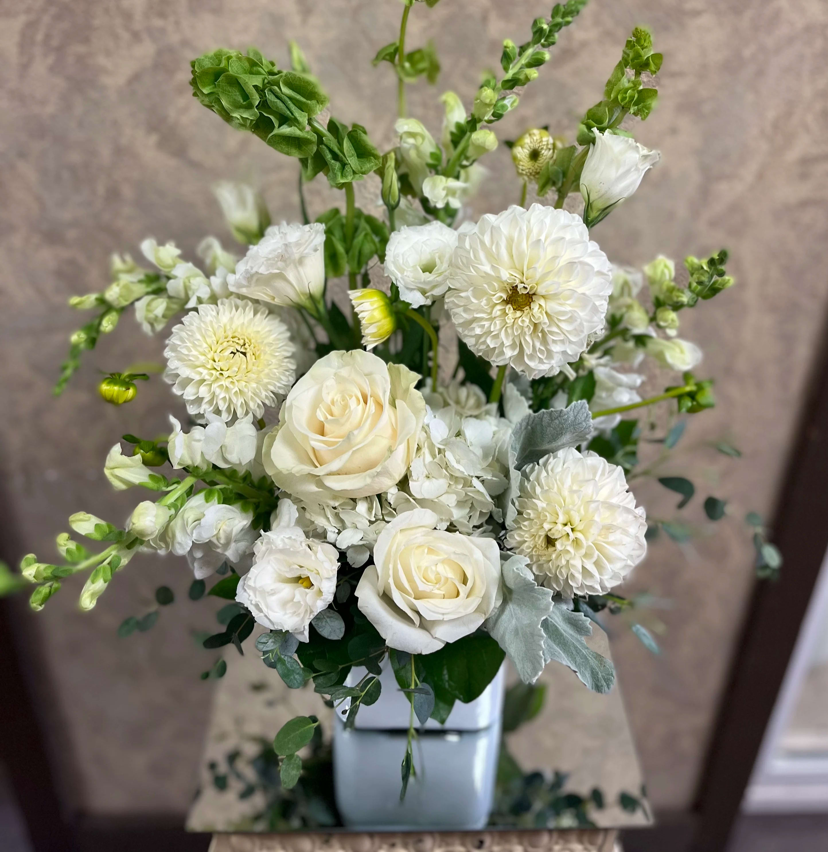 Above the Clouds - Recipe: All-white Snapdragons, Seasonal Dahlias, Roses, Lisianthus, Dusty Miller, and Eucalyptus in a short vase. Seasonal: Dahlias (mid-summer to autumn) Design View: Symmetric Front Facing View Photo shown: Standard 