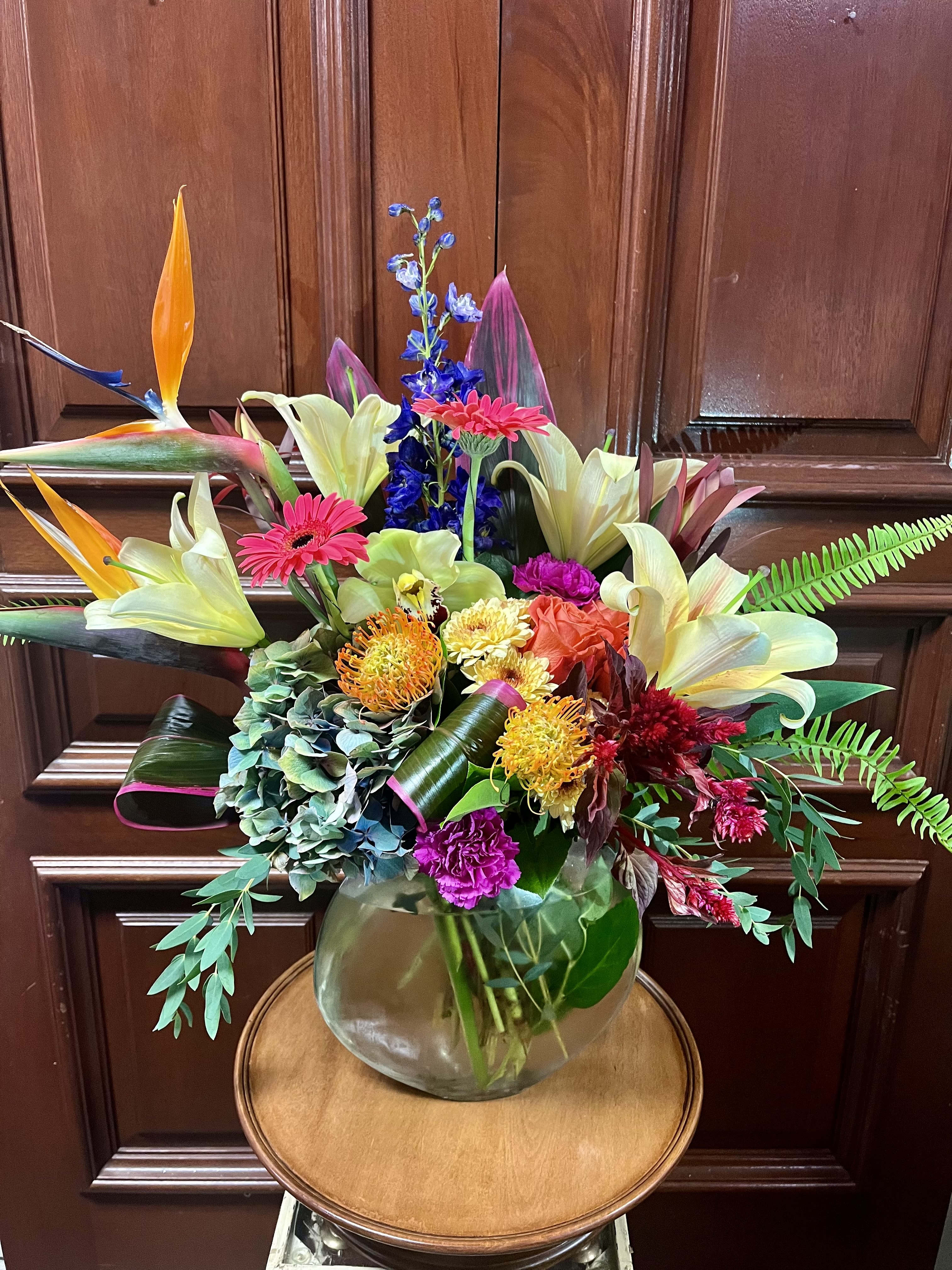 Tropical Queen NEW - Recipe: Yellow lilies, Blue Delphinium, Orange Roses, Birds of Paradise, Pincushion Protea, Green Cymbidium Orchids and more in a round vase. Seasonal: All year round Orientation: Front Facing View Photo shown: Standard