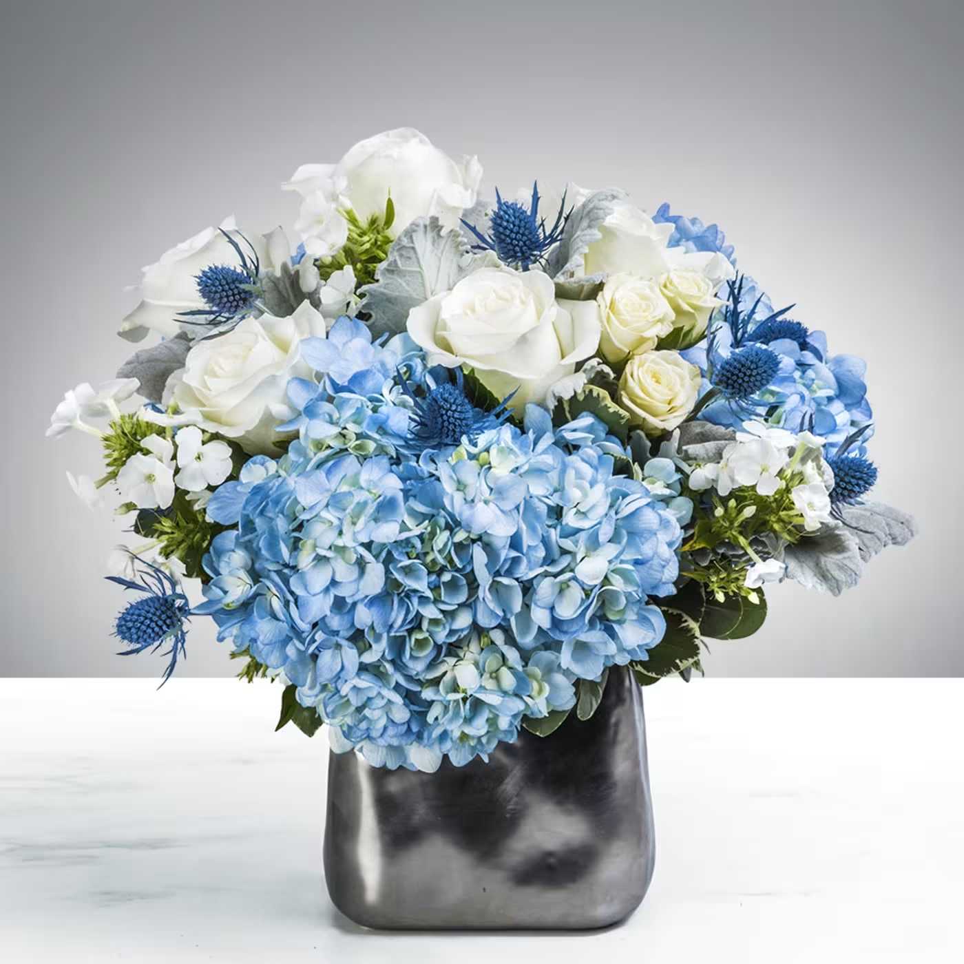 Hydrangea Lights - Beautiful arrangement in a clear square vase with blue hydrangeas, white roses, spray roses, thistle, and greenery. Vase measures approximately 5&quot;H.   
