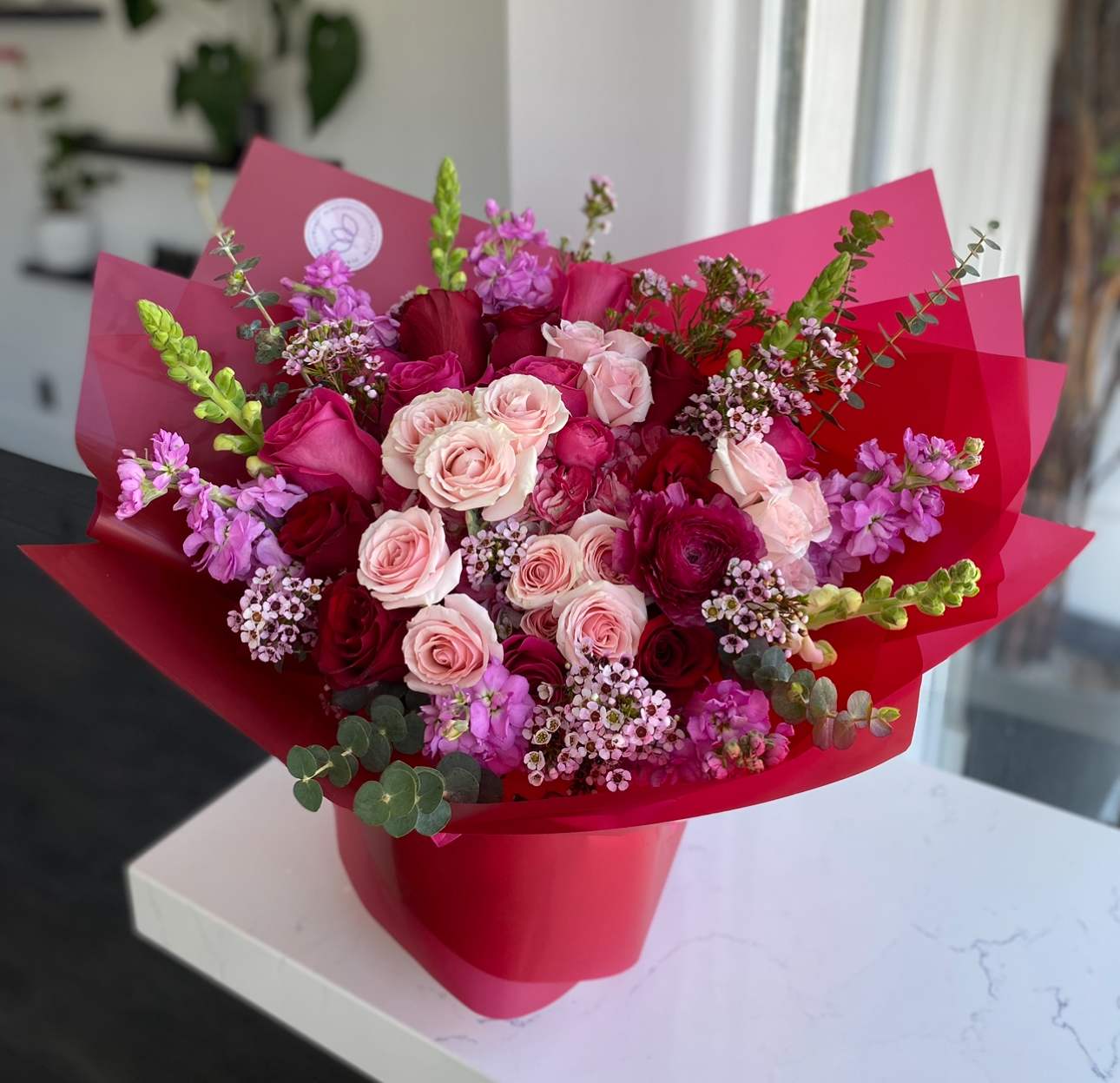 Bouquet With Love - Radiant and full of romance, the Bouquet With Love is a lush blend of premium roses in soft blush, vibrant red, and deep fuchsia tones, beautifully accented with delicate seasonal blooms and greenery. Wrapped in bold crimson paper, it makes a statement of passion and tenderness all in one. Perfect for those unforgettable moments when words aren’t enough.    •    Premium roses in blush, red, and fuchsia shades     •    Accented with seasonal fillers and fresh greenery     •    Wrapped in bold red paper with ribbon finish     •    Symbolizes deep love, passion, and devotion     •    Perfect for anniversaries, Valentine’s Day, or any romantic surprise  VASE NOT INCLUDED