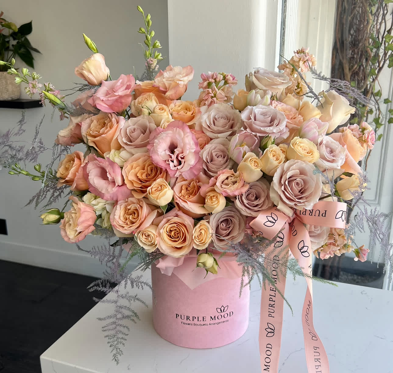 Charming Box - Standard - Container size D6.75'x H7.5' Deluxe - Container size D8.25'x H9.5'  The Charming Box lives up to its name — soft, romantic, and endlessly elegant. Designed with a luxurious blend of blush, peach, champagne, and pastel pink roses, paired with delicate lisianthus and airy greenery, this bouquet radiates grace and sophistication. Arranged in a velvet hatbox, it’s the perfect choice for expressing admiration, romance, or heartfelt celebration.    •    Premium roses and lisianthus in pastel tones     •    Blush, peach, and champagne hues for a timeless look     •    Accented with soft greenery and textures for elegance     •    Presented in a velvet hatbox with ribbon detail     •    Ideal for anniversaries, romantic gestures, or special celebrations