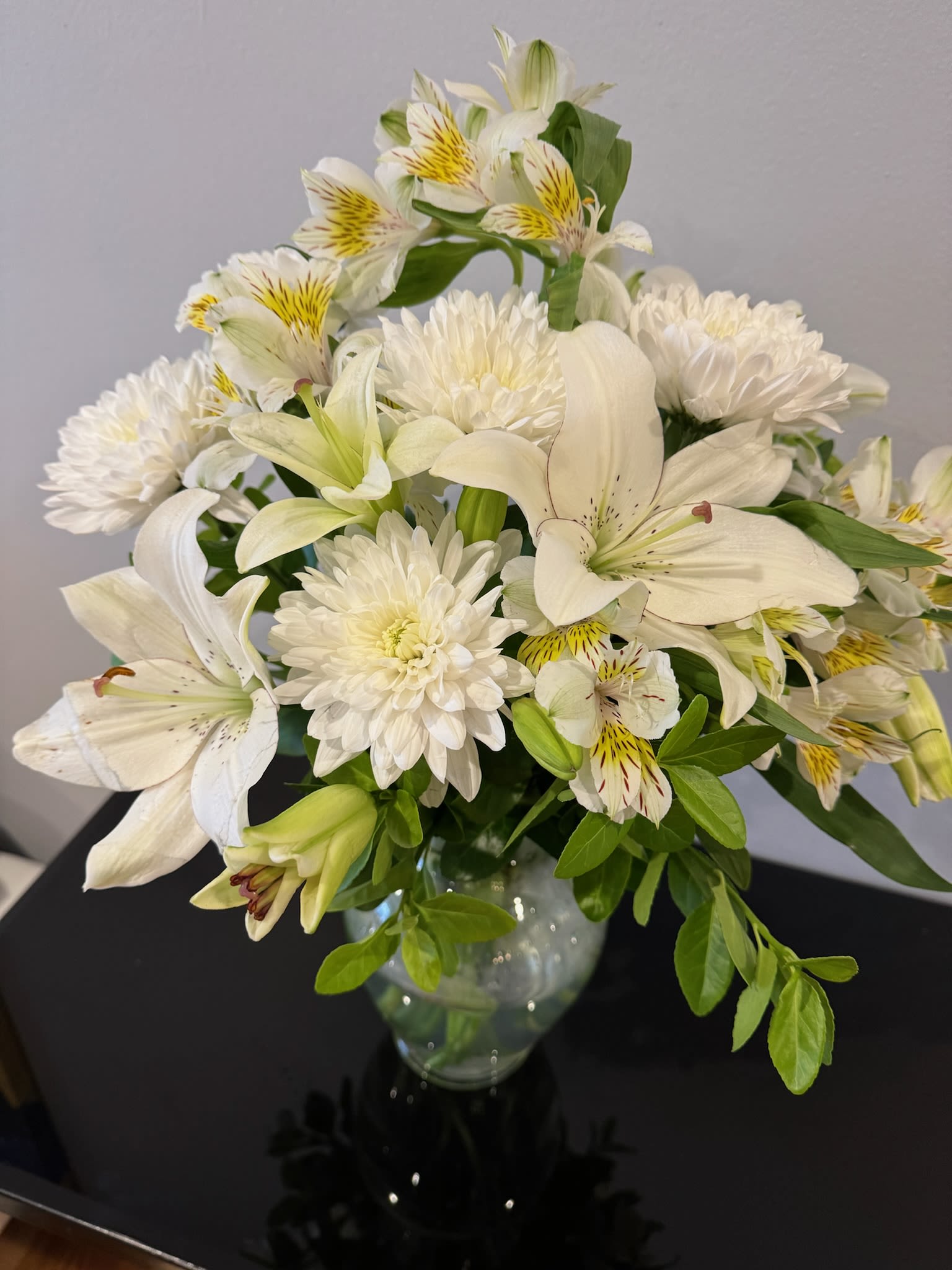 Eternal Peace Sympathy Bouquet - A sympathy arrangement featuring White Mums, White Lilies, Alstroemeria, and assorted greens would convey a serene and comforting sentiment. The combination of white flowers symbolizes purity, peace, and reverence, which are fitting for expressing condolences and sympathy. The lush greens add texture and depth to the arrangement, enhancing its natural beauty and providing a sense of tranquility. Overall, it's a thoughtful choice to offer solace and support during a difficult time.  White Mums White Lily Alstroemeria  Height 22 inches Width 14 inches