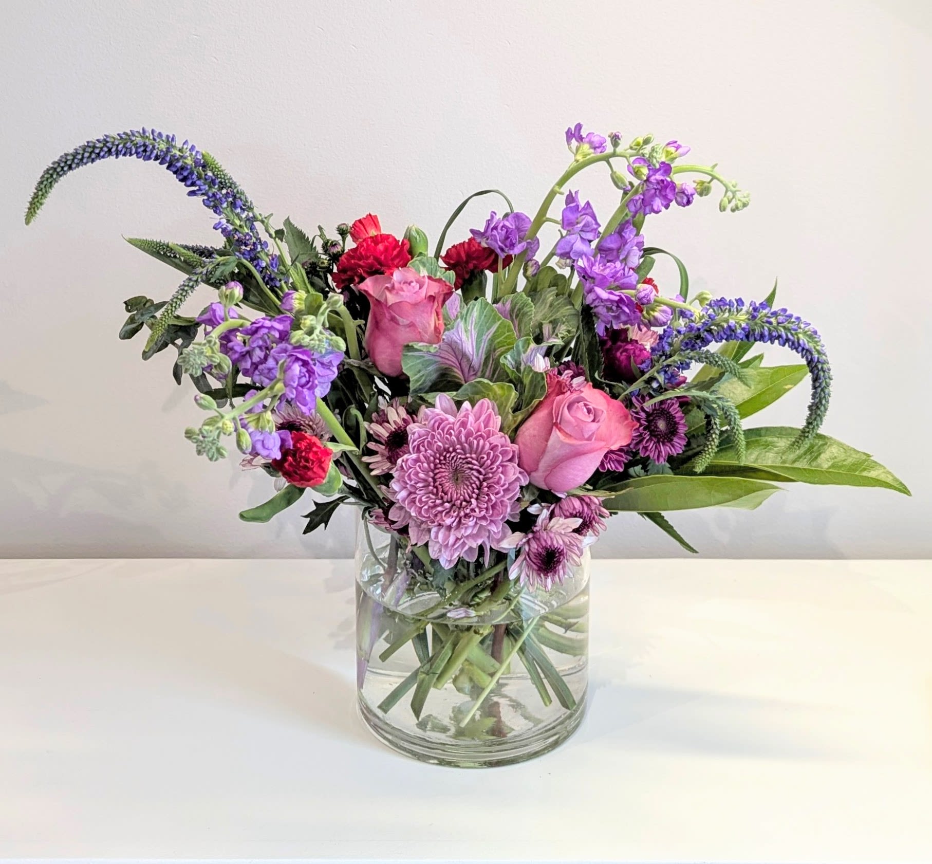 Sugar Magnolia Sugar Magnolia - A lush floral bouquet using blooms in lavender, purples, green & a pop of red. This modern & beautiful arrangement is designed in a classic clear glass cylinder & is the perfect gift for all occasions. Measures approx. 12" high & 15" wide Orientation: All around