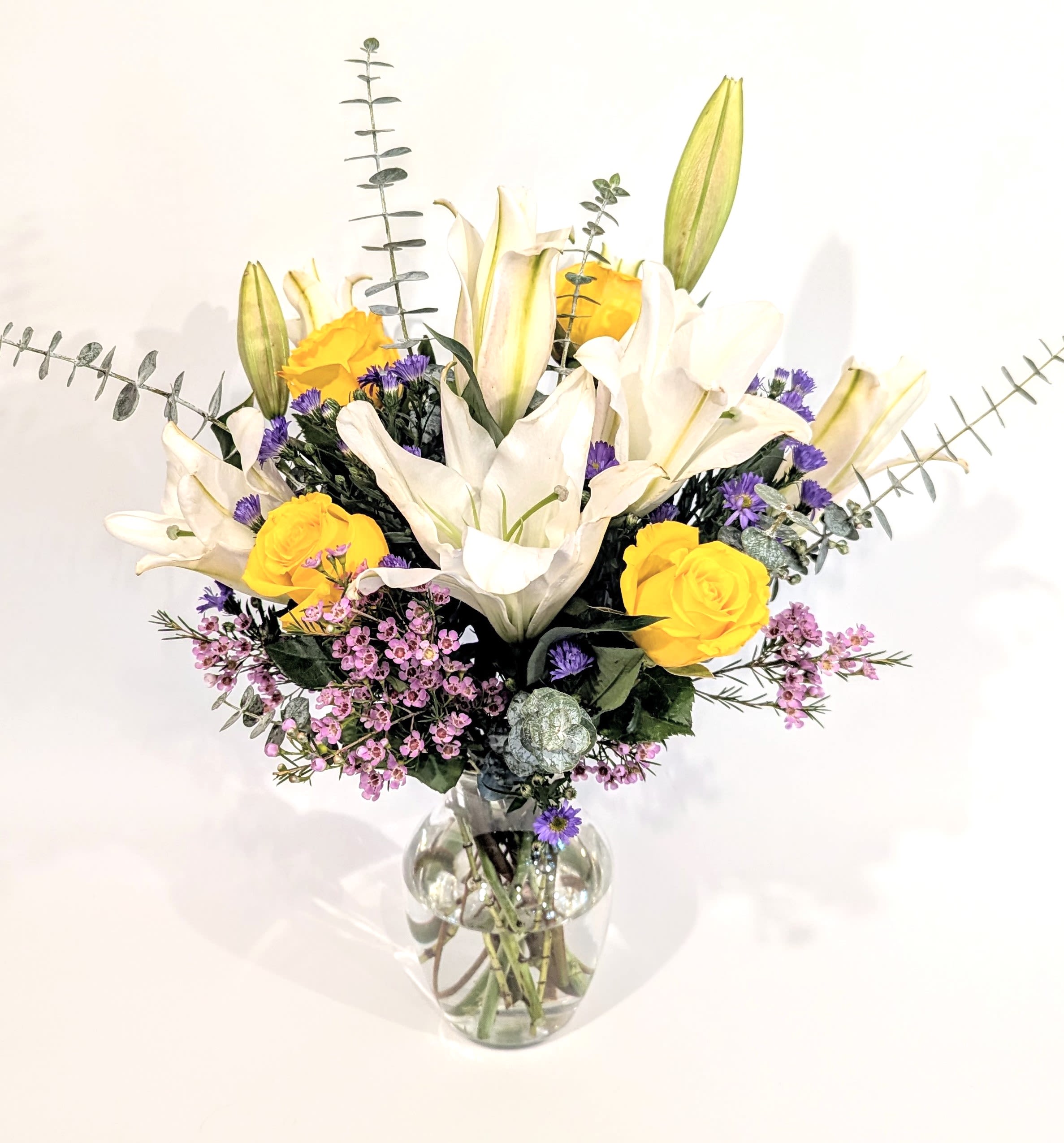 Friendship Bouquet  - A lovely and fragrant bouquet full of white lilies, lavender wax flower, purple monte casino, yellow roses and spiral eucalyptus. Arranged with a soft palette of seasonal blooms and delivered in a clear glass rose vase. The arrangement is suitable for all occasions especially just because and get well.  Measures approx. 23&quot; tall and 19&quot; wide Orientation: All around
