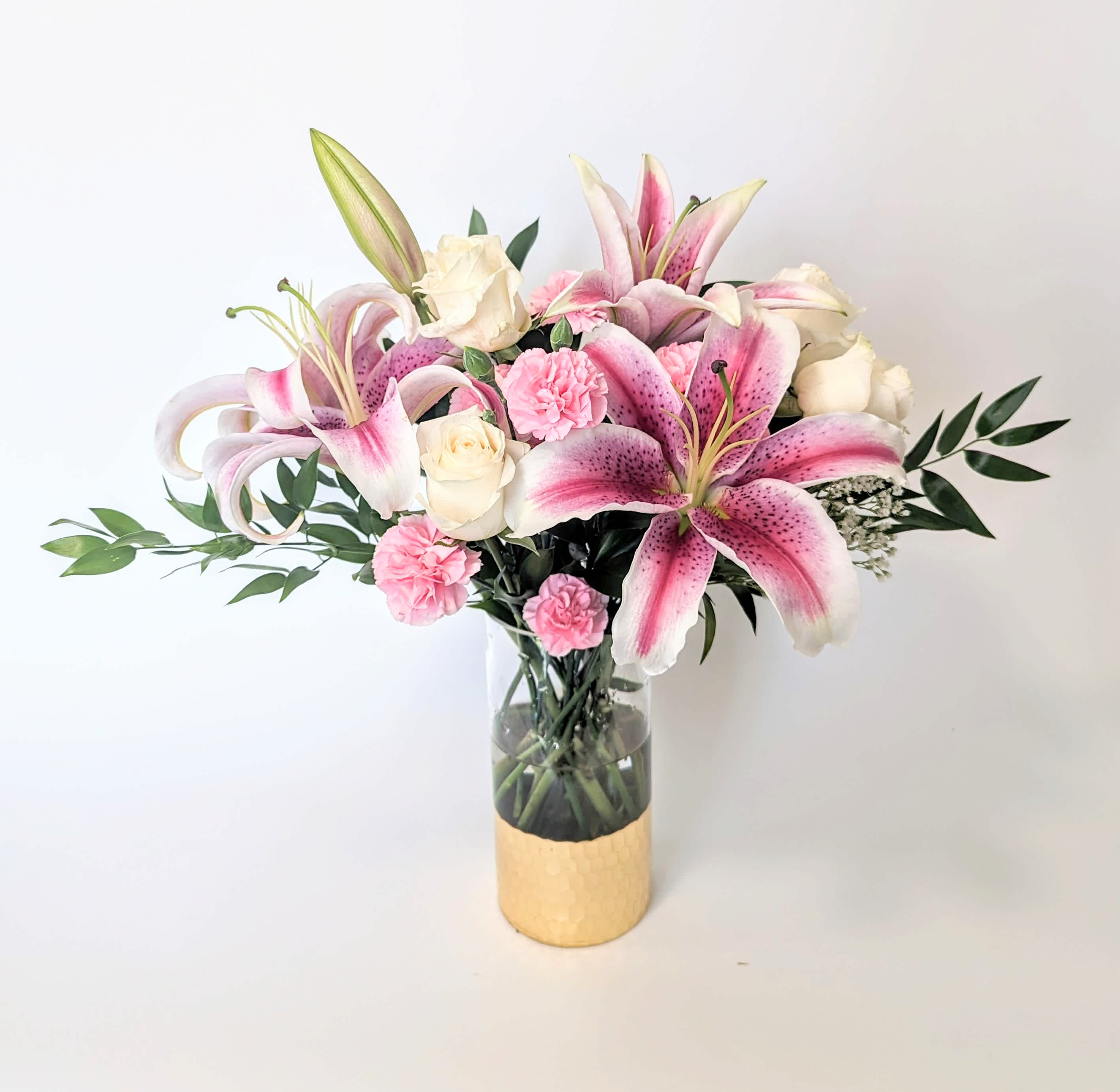 Elegant Enchantment - Emanating an aura of timeless elegance and delicate beauty, our exquisite arrangement features a captivating ensemble of oriental lilies, their graceful petals unfurling in a mesmerizing display of purity and sophistication. Amidst this floral symphony, pristine white roses stand as symbols of purity and reverence, their velvety blooms exuding an ethereal charm that captures the essence of love and admiration.  Complementing this celestial palette, clusters of pink mini carnations add a playful touch of warmth and affection, their dainty blossoms infusing the arrangement with a sense of joy and sweetness tucked among fragrant eucalyptus greens. Delivered in a classic gold-dipped glass cylinder vase, this arrangement becomes a work of art, marrying contemporary opulence with natural splendor.  Measures 18&quot; high and 18&quot; wide