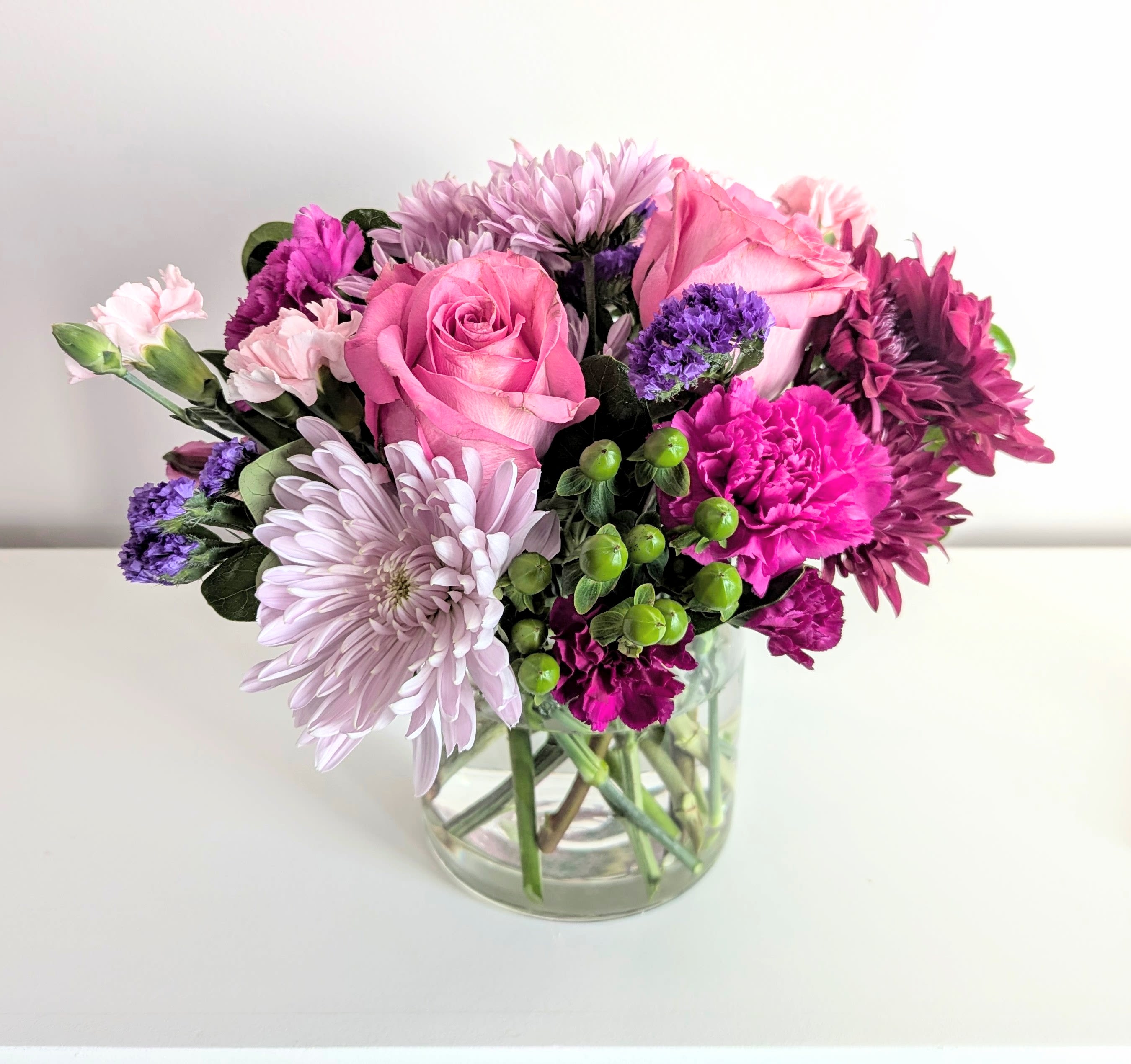 Strawberry Fields - This bouquet is bursting with lots of juicy blooms, like hot pink roses, burgundy &amp; lavender mums, pink carnations, alstroemeria &amp; greenery. Arranged low &amp; compact in a clear glass cylinder vase. This arrangement is suitable for any special occasion.   Measures approx. 13&quot; wide &amp; 11&quot; tall Orientation: All around 