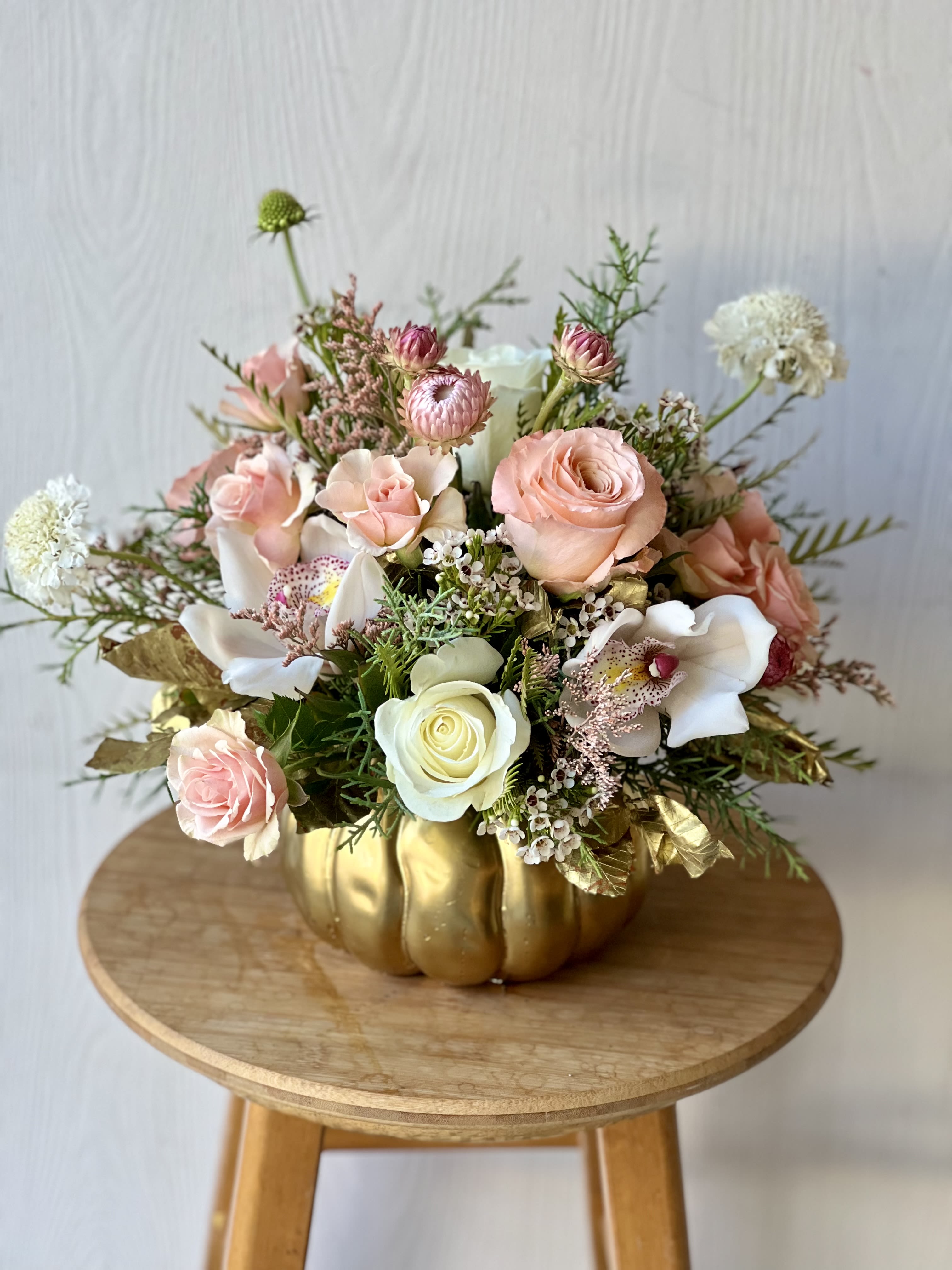 EC123 Golden Pumpkin - Celebrate the beauty of fall with our exquisite Golden Pumpkin Floral Arrangement. Designed in an elegant gold pumpkin vase, this stunning centerpiece showcases a harmonious blend of peach roses, peach spray roses, and white roses, perfectly paired with luxurious white cymbidium orchids. Accented with delicate white wax flowers and lush seasonal greenery, this arrangement radiates warmth and sophistication.  Ideal for Thanksgiving tablescapes, fall décor, or as a thoughtful gift, the Golden Pumpkin adds a touch of elegance to any setting. Order now for same-day delivery and make your holiday season truly memorable!