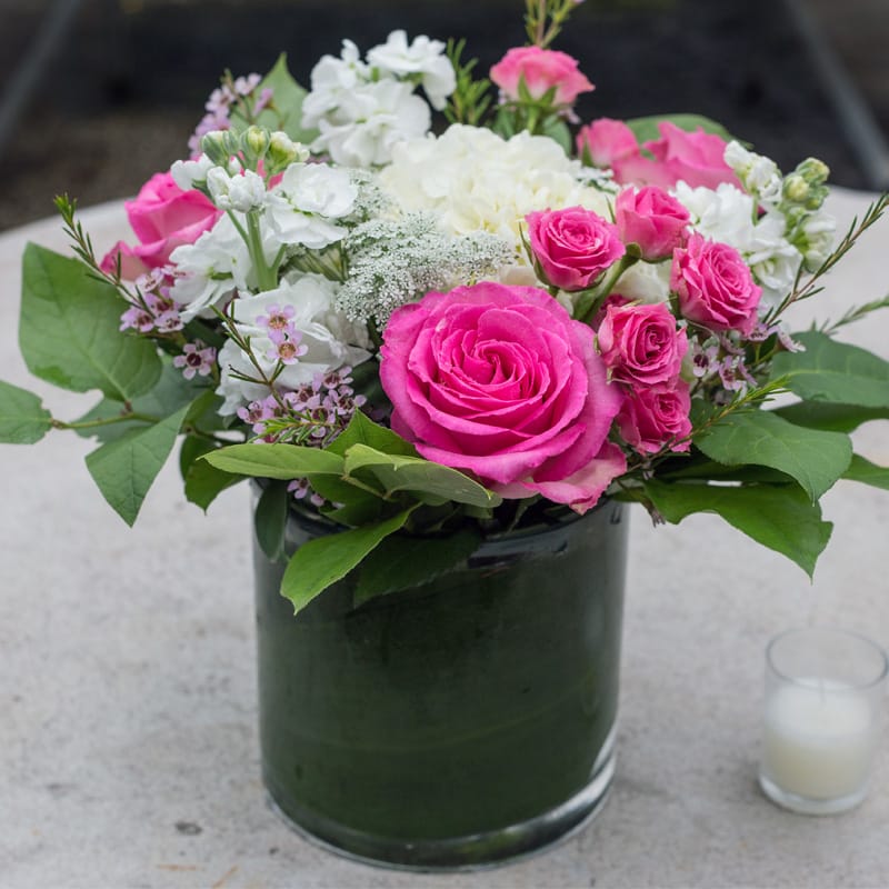 Pink Lady - Hydrangeas, Queen Ann's lace, stock, spray roses, wax flower, lemon leaf, and a sweet unique rose in a cylindrical glass vase. 