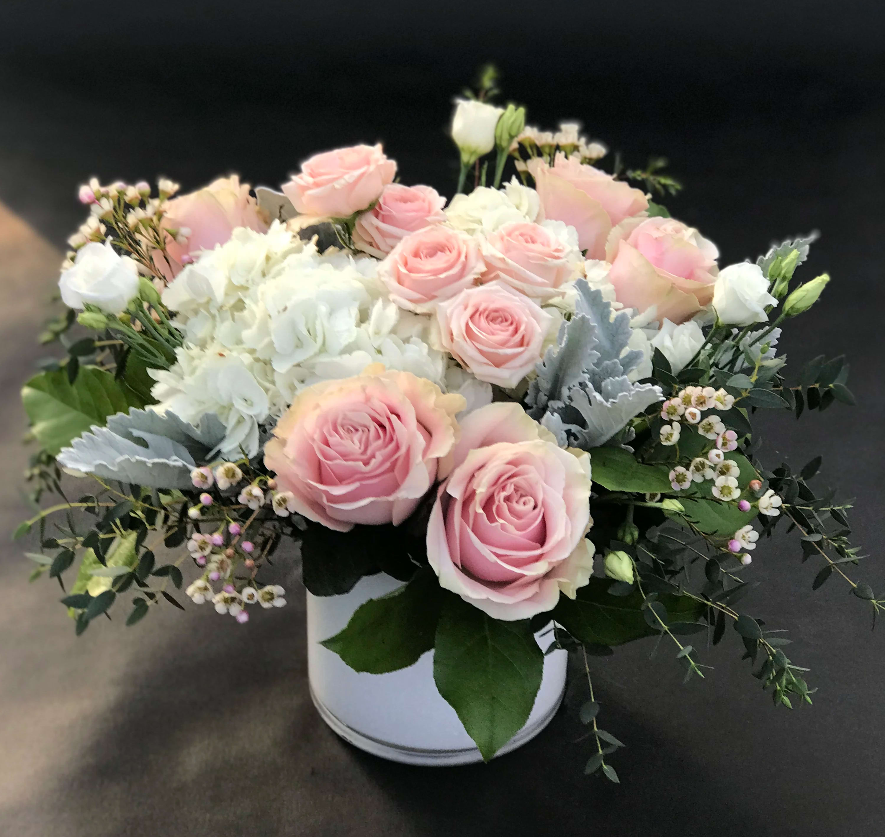 Pink Sweetheart  - Pink Mondial roses, pink spray roses, white hydrangea with contrasting cool green Dusty Miller and springs of delicate wax flower in an elegant white glass cylinder. 