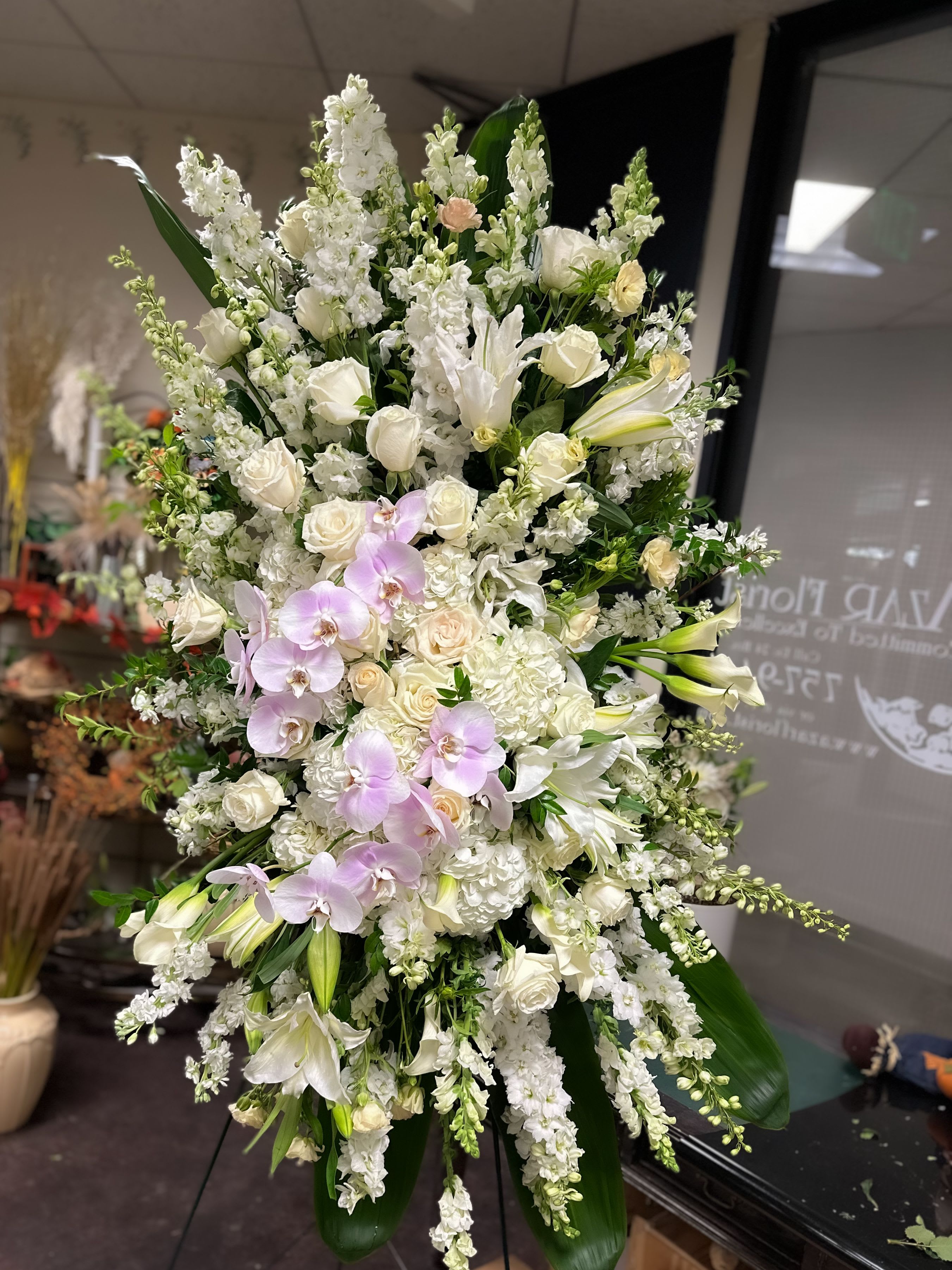 Pacific View Elegance Tribute - An awe-inspiring 8-foot standing spray created to honor a life with grace and serenity. Designed in all white, this striking piece blends classic lilies, lush roses, and tall delphinium with cascading phalaenopsis orchids that flow softly through the arrangement. The towering height and pure palette create a sense of peace, making it a fitting tribute for services at Pacific View and throughout Orange County.
