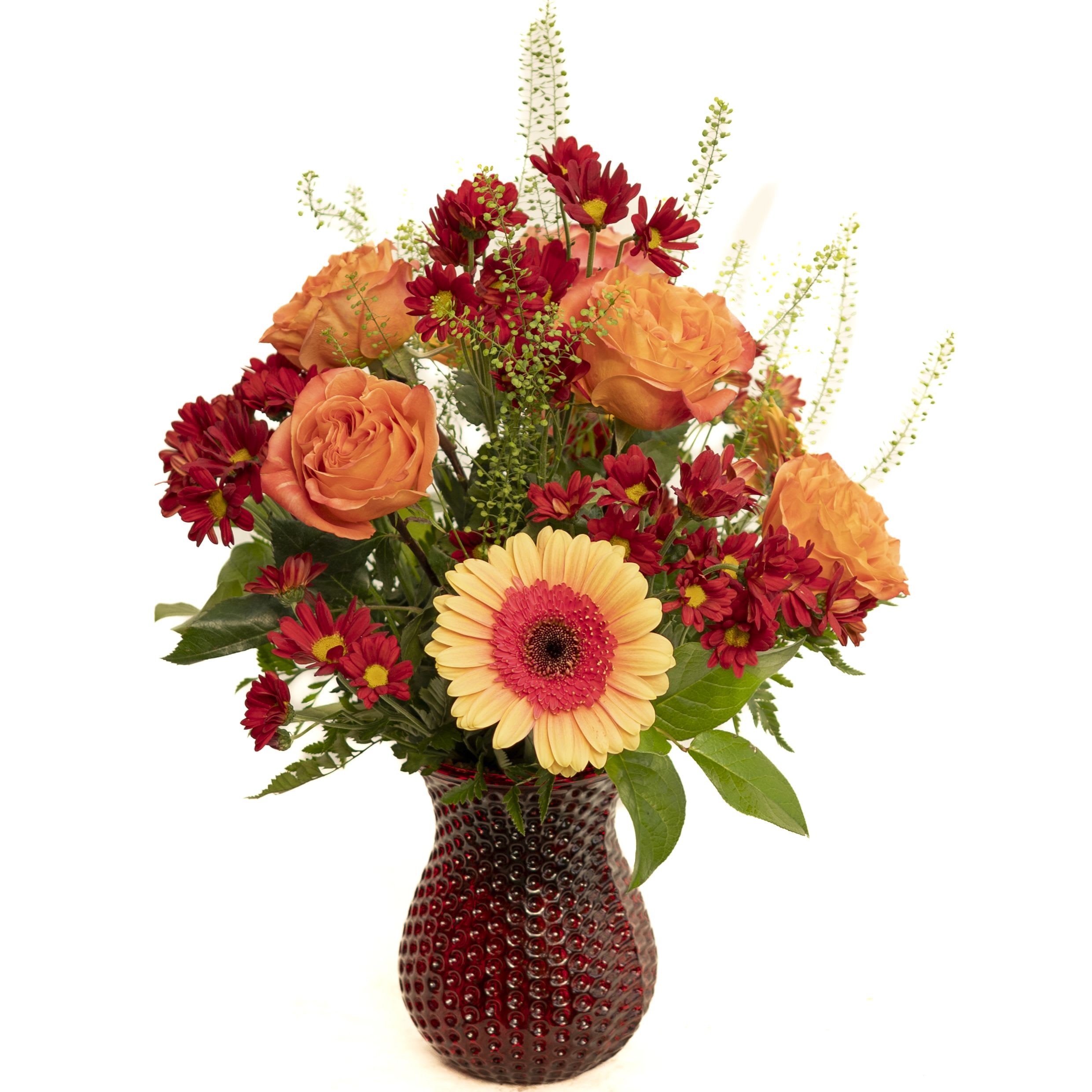 Autumn Glow (red vase) Autumn Glow (red vase) - Bright and cheerful, the Autumn Radiance Bouquet features a warm blend of orange roses, deep red daisy poms, and a striking peach gerbera daisy accented with fresh greenery. Arranged in a textured red glass vase, this design brings a cozy seasonal charm, perfect for adding a burst of color to any room or gifting to someone special.