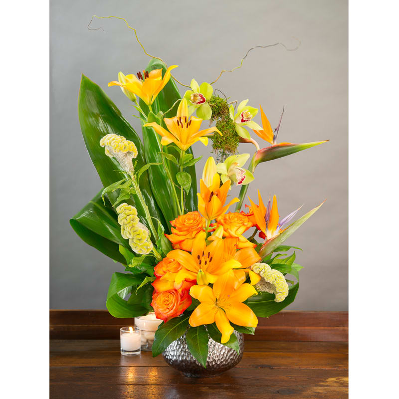 ORANGE CRUSH - LILIES, BIRDS OF PARADISE, ROSES, TROPICAL GREENS, CYMBIDIUM ORCHIDS IN A SILVER CERAMIC CONTAINER