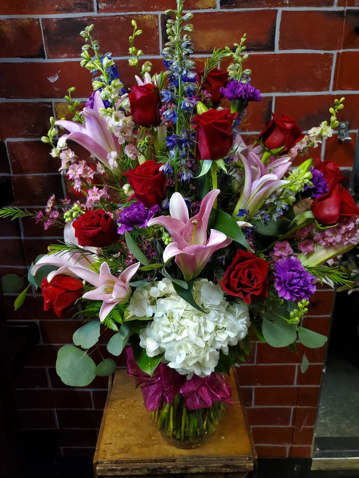 He Luvs Me - Celebrate love and beauty with our He Luvs Me Bouquet, featuring a stunning mix of a dozen classic red roses, soft pink lilies, pristine white hydrangea, delicate pink larkspur, striking blue delphinium, and rich purple carnations, all tied together with a pink bow. This colorful, elegant arrangement is perfect for anniversaries, birthdays, or any moment when you want to make someone feel truly special.  This is a one sided bouquet.  Each bloom is hand-selected for freshness and expertly arranged with care to create a breathtaking display. Delivered by Adorkable Flowers and Gifts, the Romance in Bloom Bouquet is a vibrant and heartfelt gift that speaks directly from the heart. * Some stages of flower life may vary. (Example: Lilies may be more open than imaged.)  Flower shortages and product availability means that some substitutions will apply. Value will always be filled.  This bouquet might include eucalyptus amongst the greenery. It may be toxic to pets including but not limited to Cats and Dogs if digested.
