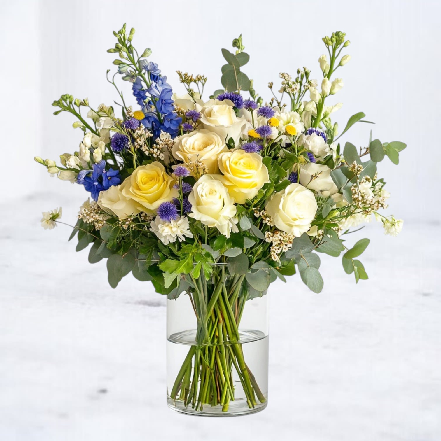 Blue Skies & Buttercream Blue Skies & Buttercream - Lift their spirits with Blue Skies & Buttercream — a soft, serene bouquet featuring pale yellow roses, white lisianthus, and sky-blue delphinium accented by touches of deep blue cornflower and airy filler textures. Designed in a low-profile glass vase, this gentle arrangement is equal parts cozy and refreshing. Perfect for Get Well wishes, recovery gifts, or simply sending a moment of peace and beauty. Hand-delivered with same-day service in Houston, Magnolia, Montgomery, Conroe, The Woodlands, and Spring.