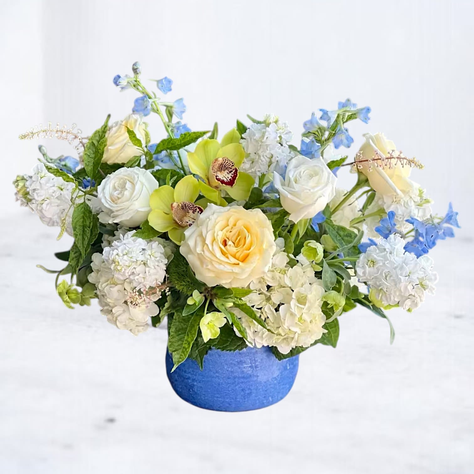 Bluebell Reverence Bluebell Reverence - Elegant and serene, the Bluebell Reverence arrangement is a sophisticated floral tribute perfect for expressing sympathy or sending peaceful thoughts. Designed in a cobalt blue vase, this lush design features creamy roses, white stock, hydrangea, light blue delphinium, and exotic green cymbidium orchids. The gentle color palette offers comfort and grace, making it ideal for a sympathy gesture, memorial service, or hospital delivery. Available for same-day delivery in Houston, The Woodlands, Magnolia, and surrounding areas.