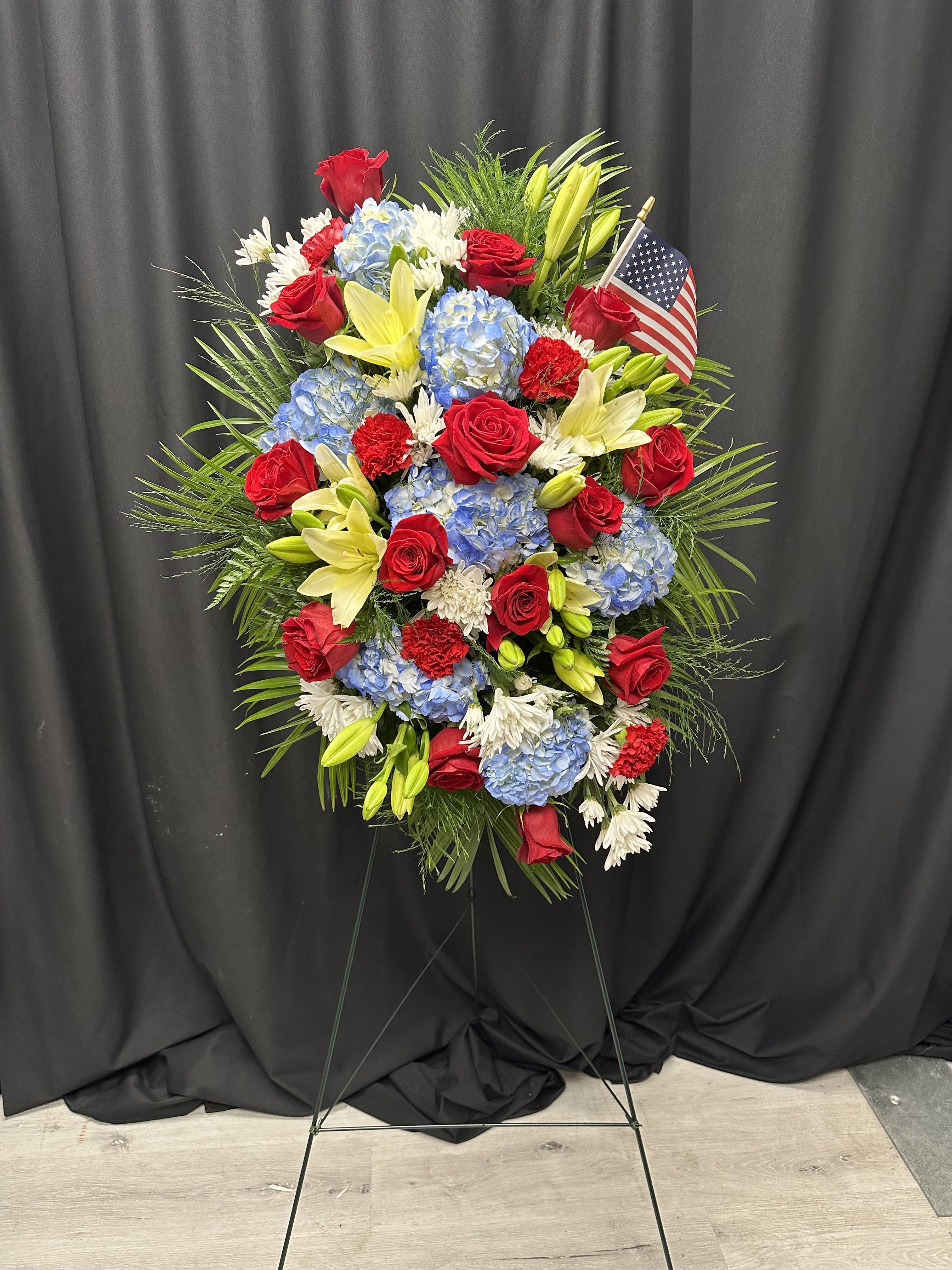 Honored Patriot Honored Patriot - Honor your greatest patriot with a majestic standing spray adorned with vibrant red gerbera daisies, rich red carnations, pure white mums, fragrant white stock, and majestic blue delphinium. A customizable blue bow adds a personal touch, while the presence of the American flag accentuates this magnificent tribute. May this symbol of love and respect accompany your farewell with dignity and pride, commemorating a life devoted to country and duty.