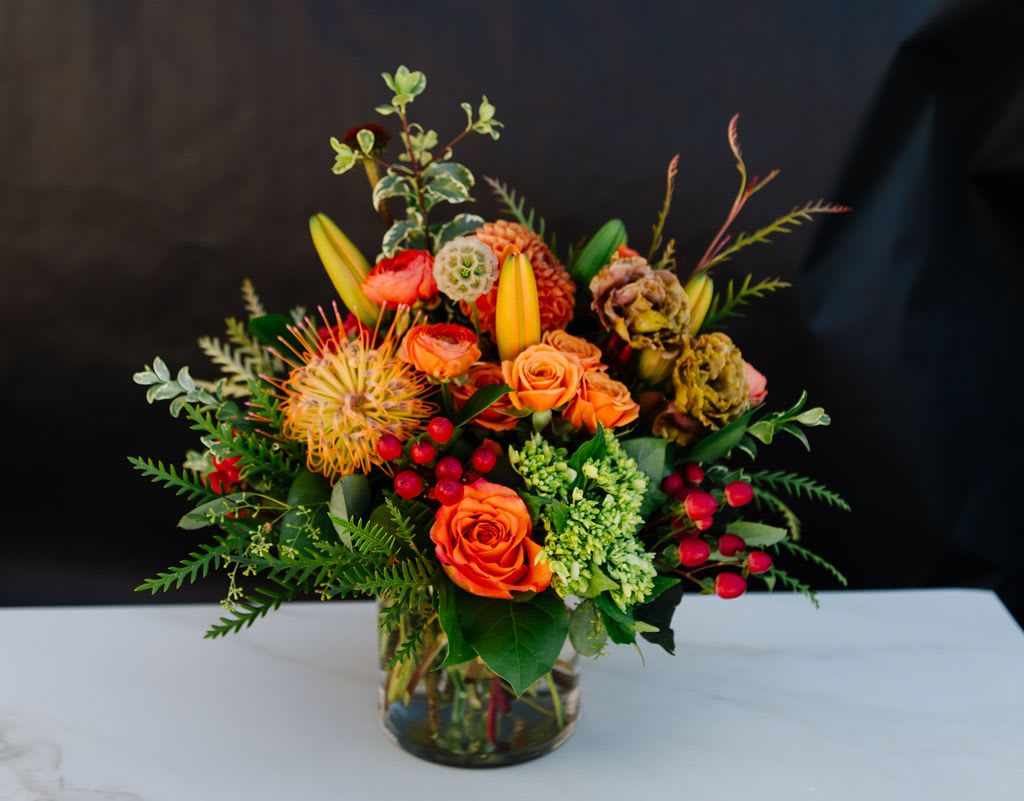 Amber Blooms - Bring the warmth of fall into any space with our Amber Blooms arrangement. Featuring vibrant orange roses, golden pincushion protea, lilies, and lush greenery accented with red hypericum berries, this design bursts with color and texture. Perfect as a seasonal centerpiece, a thoughtful gift, or a way to brighten someone’s day, Amber Blooms captures the rich beauty of nature in full autumn colors.