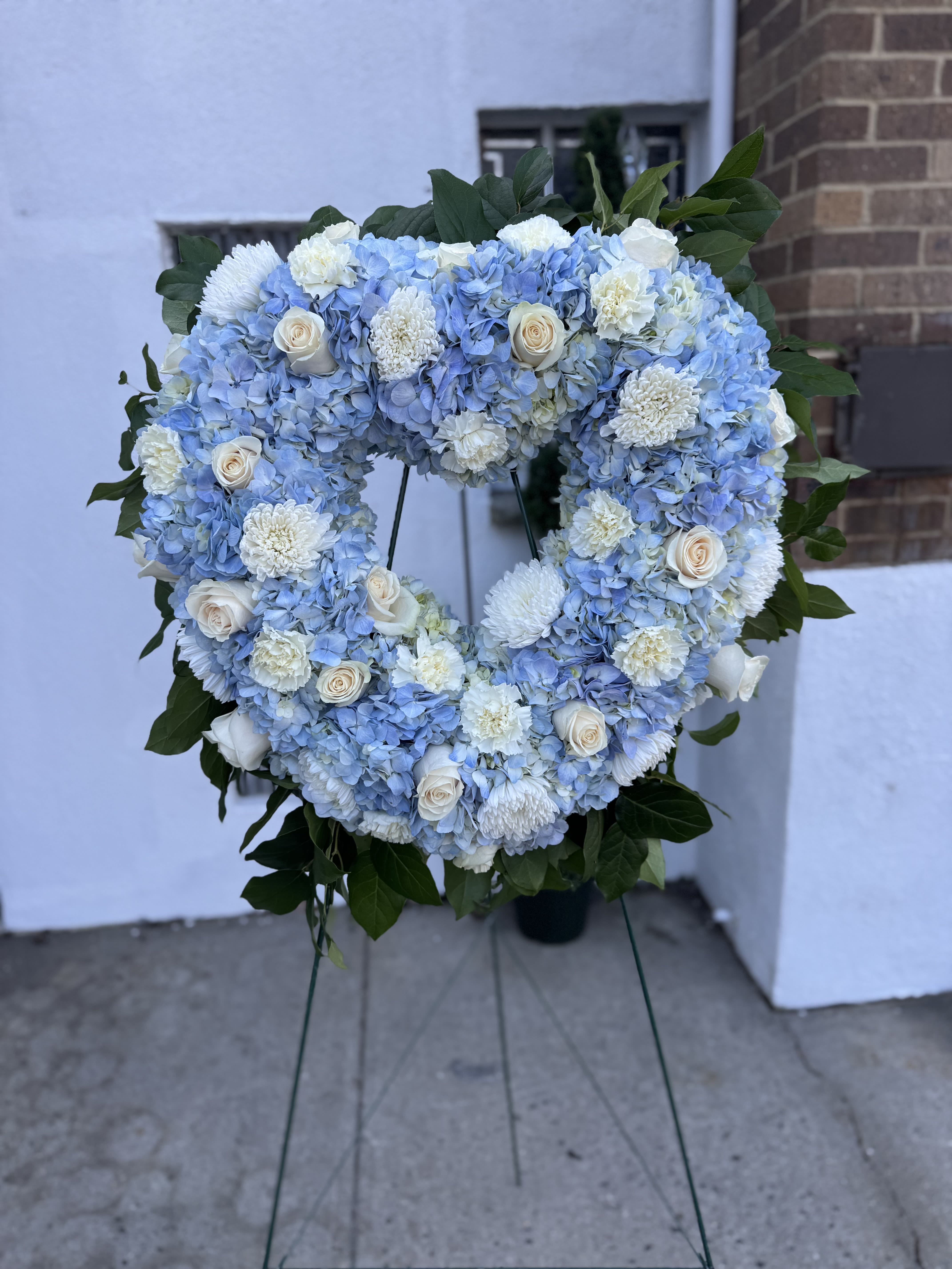 Beloved and Treasured Open Heart - A beautiful way to honor your loved one's memory. This open-heart spray is artfully arranged with blue hydrangeas, white roses, white mums, and white carnations, accented with fresh greenery for graceful and uplifting tribute.