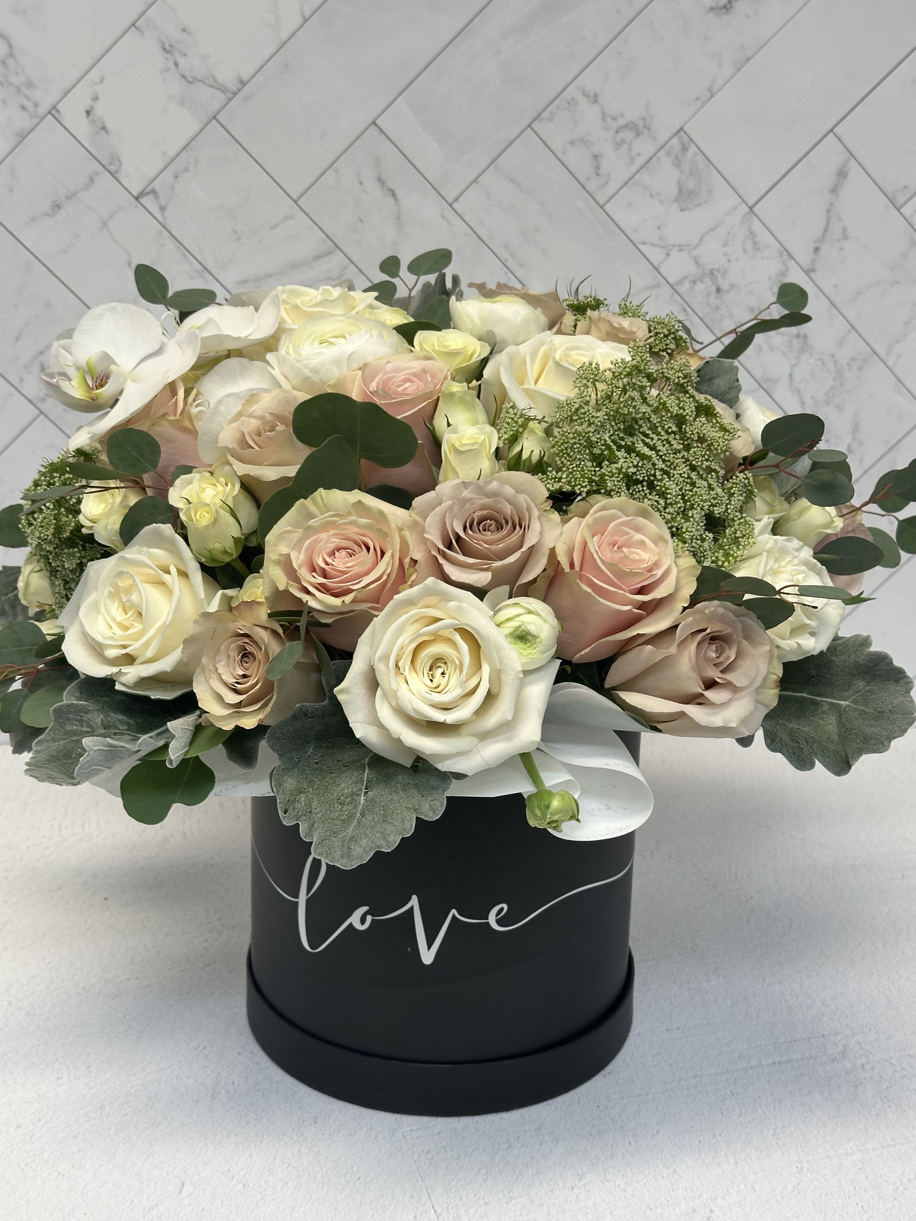 Melisa - A luxurious mix of white, light pink, and beige roses paired with elegant white ranunculus, orchids, and lush mixed greens. Designed in a stylish hat box (box color may vary based on availability)  