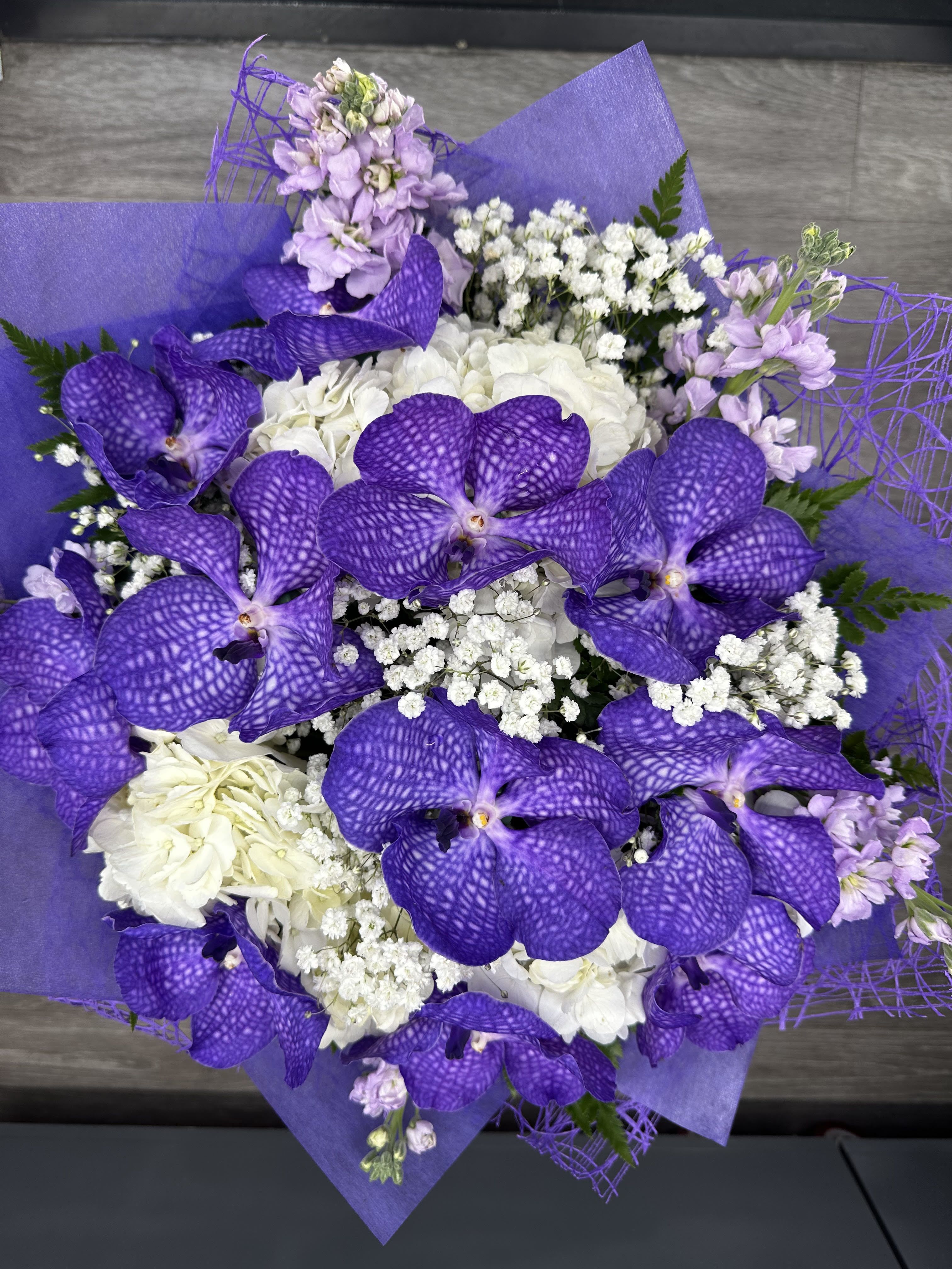 Violet  - One of our best sellers, this stunning bouquet blends shades of purple and white for a truly elegant look. Featuring exotic purple Vanda orchids, lush white hydrangeas, delicate baby's breath , and fragrant lavender stock, it's beautifully wrapped in rich purple paper. This arrangement is designed without a vase, but you may add one to your order for an extra touch of elegance. 