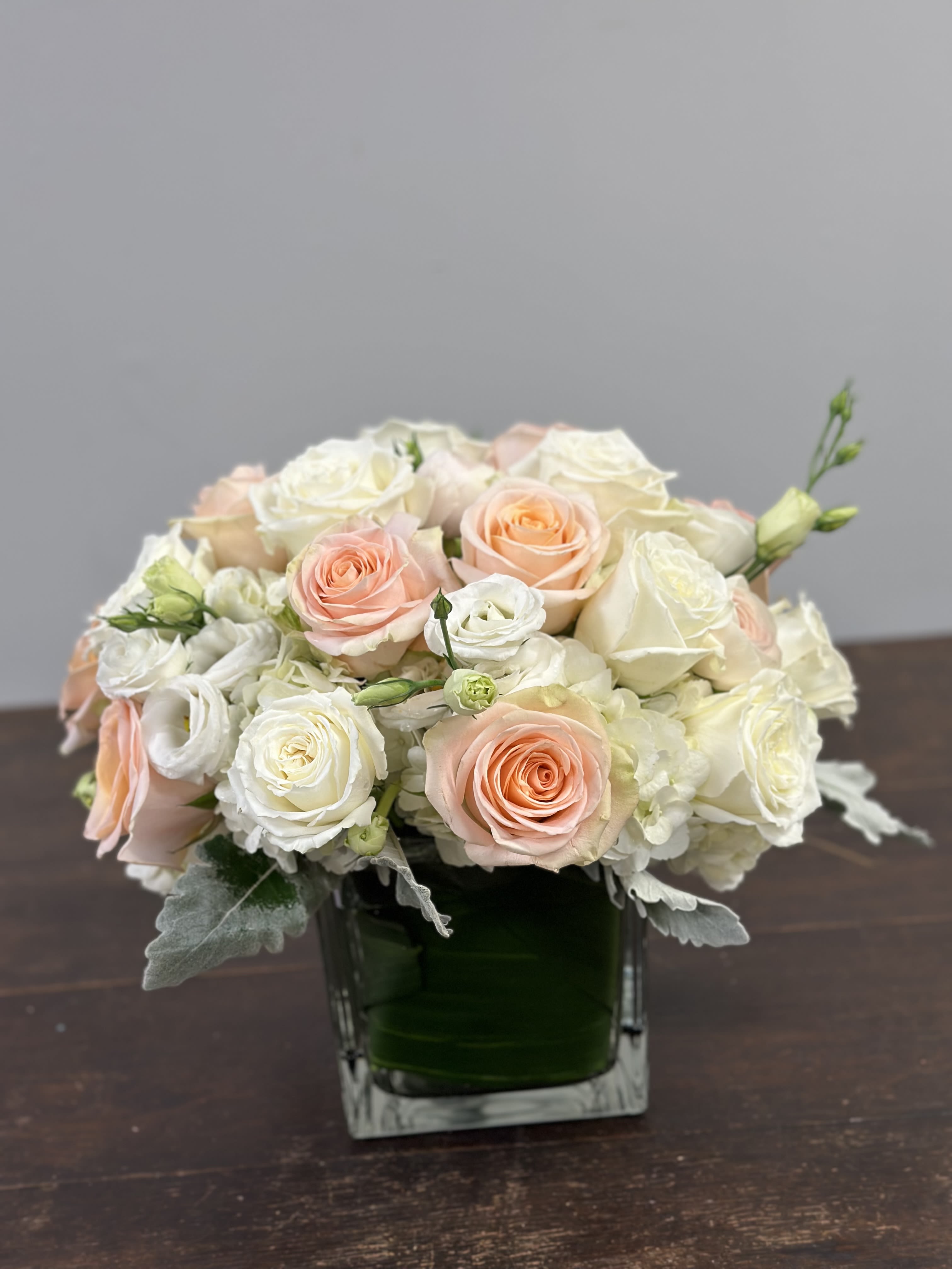 Charming Glow - A soft mix of peach and white roses with lisianthus, white hydrangeas, ranunculus, and greenery, arranged in a vase. A simple yet elegant design to brighten any room.