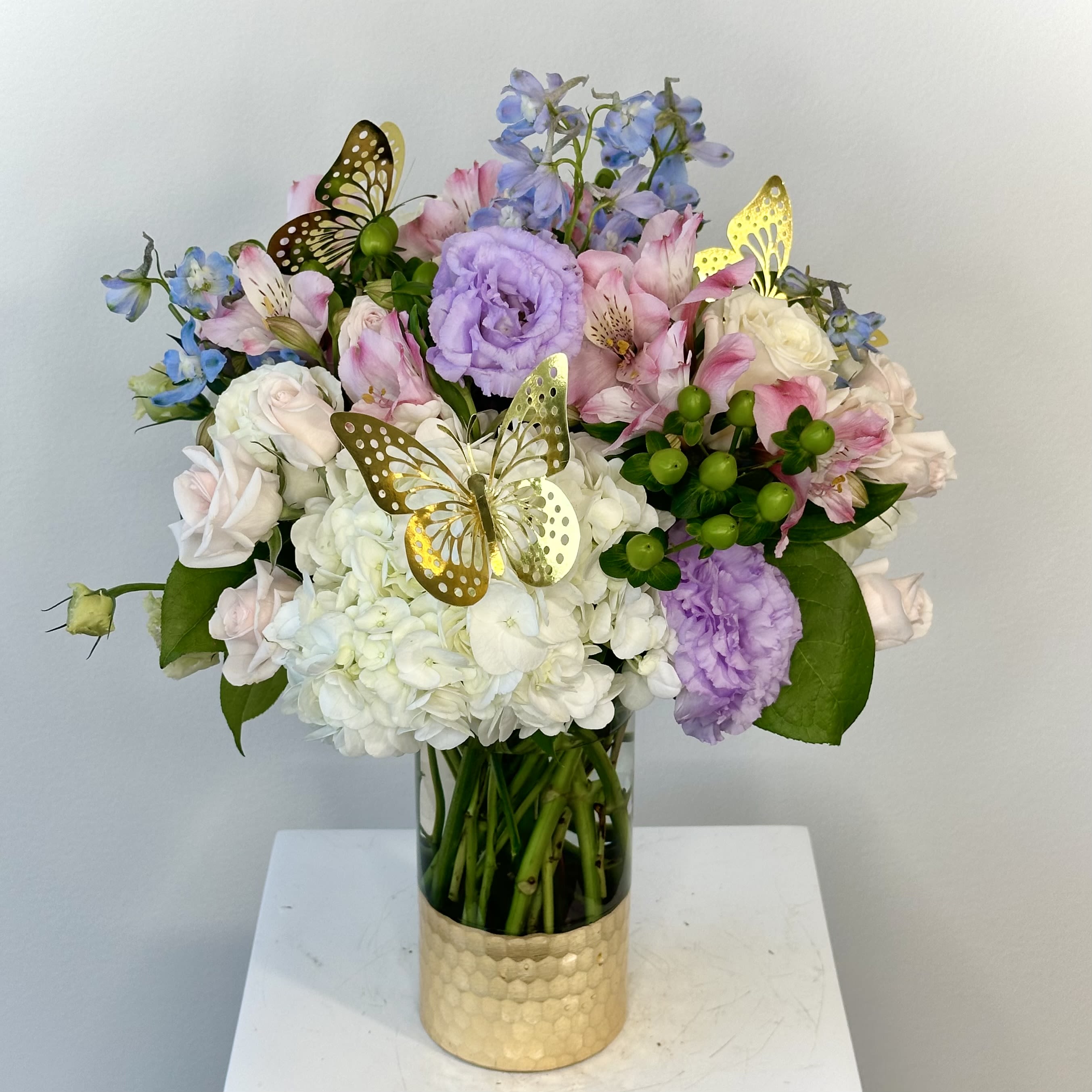 Make Me Flutter - “Make Me Flutter” is an exquisite arrangement that features gorgeous lisianthus and delphinium that compliment each other with their synonymous blue and purple hues that pair well with the pink color of alstroemeria. To tie everything together, it also includes hydrangea, roses, and hypericum berries. 