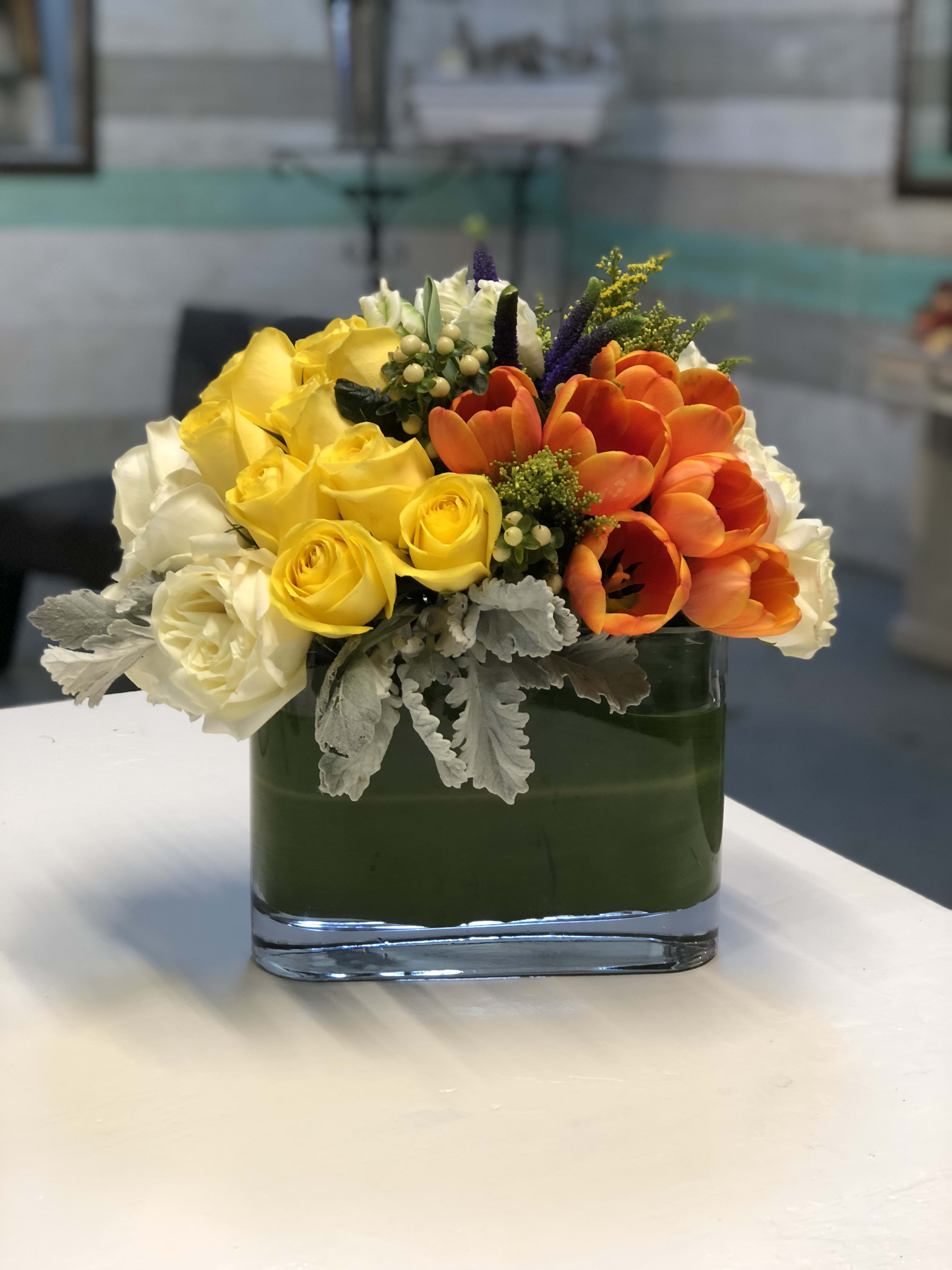 Bernal Heights Delight - A creative arrangement of papaya-orange Tulips, sunshine-yellow and white Roses, Dusty Miller, seasonal greens and berries in an ovular, tropical leaf-lined glass vase.