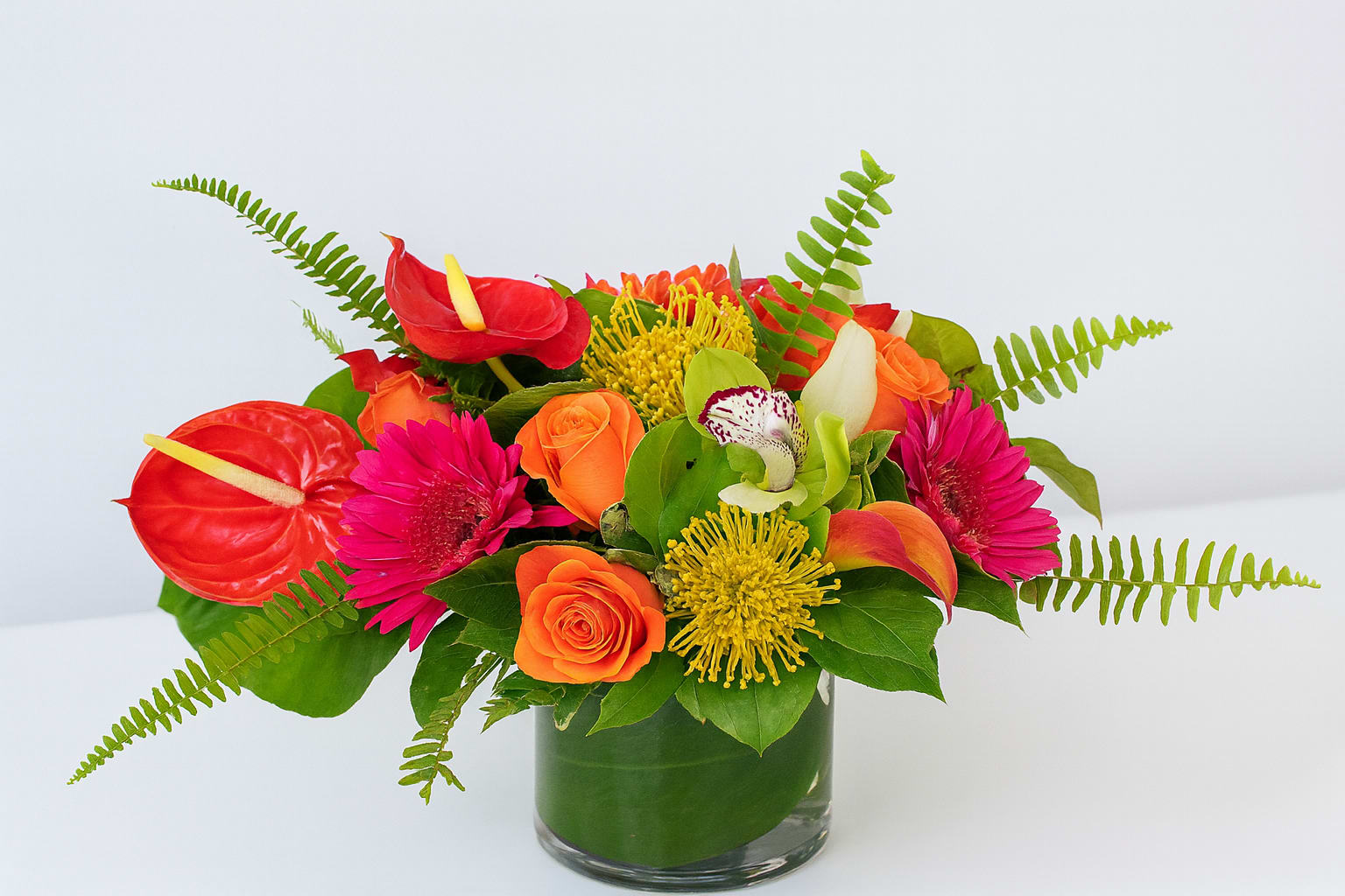 Tropical Sunset Glow Tropical Sunset Glow - Bold, exotic, and full of energy, this arrangement captures the warmth of a tropical sunset. Bright orange roses, vivid fuchsia gerbera daisies, and striking red anthuriums take center stage, while golden pincushion protea and a delicate cymbidium orchid add an unexpected touch of luxury. Fresh greenery and playful fern fronds complete the look, giving the design a lively and modern feel. Designed in a sleek glass cylinder vase, Tropical Sunset Glow is perfect for making a statement at dinner parties, celebrating special occasions, or gifting someone who loves vibrant, unforgettable flowers.