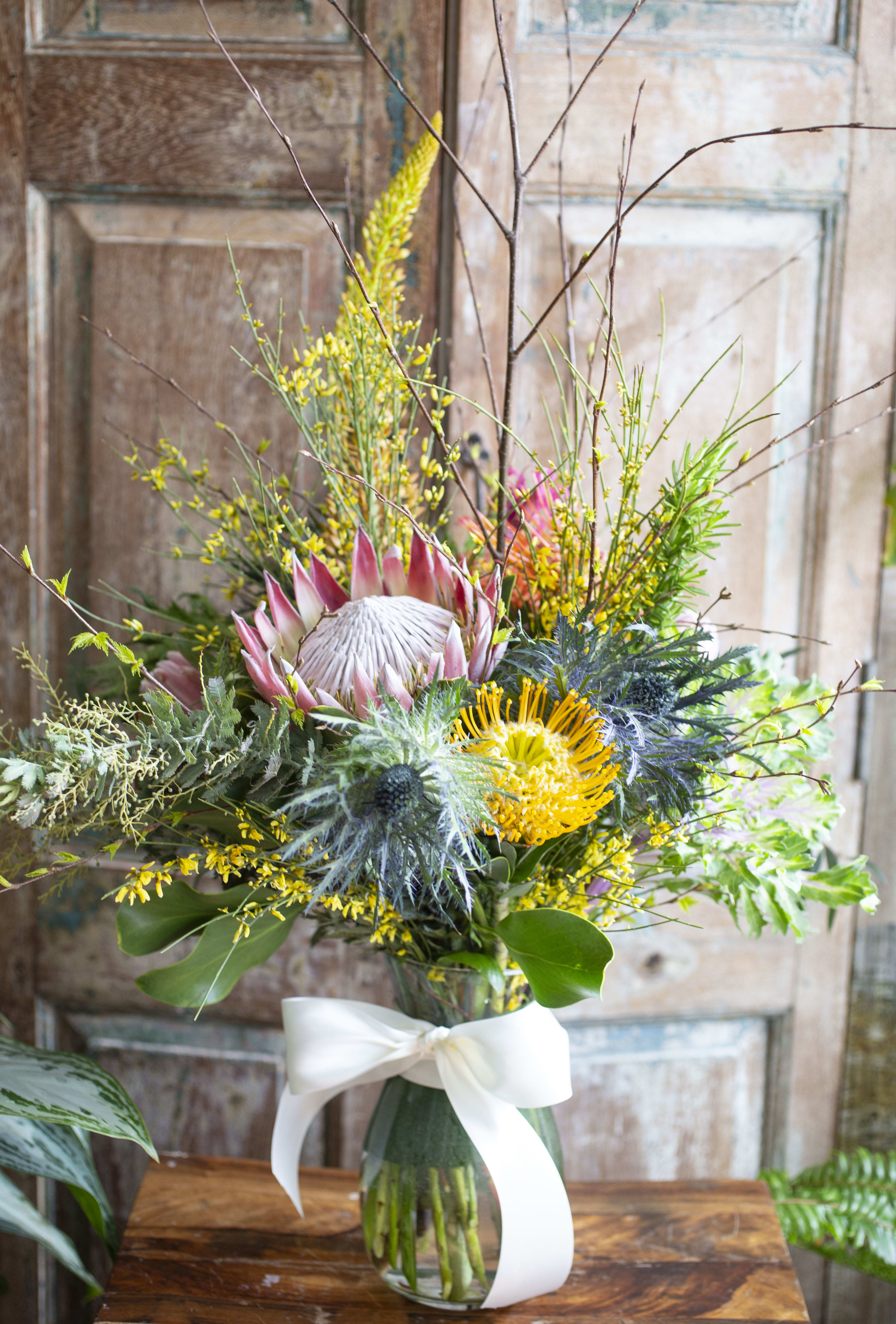 Liam - King Protea, Pin-Cushion Protea, Thistle, unique seasonal accents and foliage arranged in a clear glass vase. 