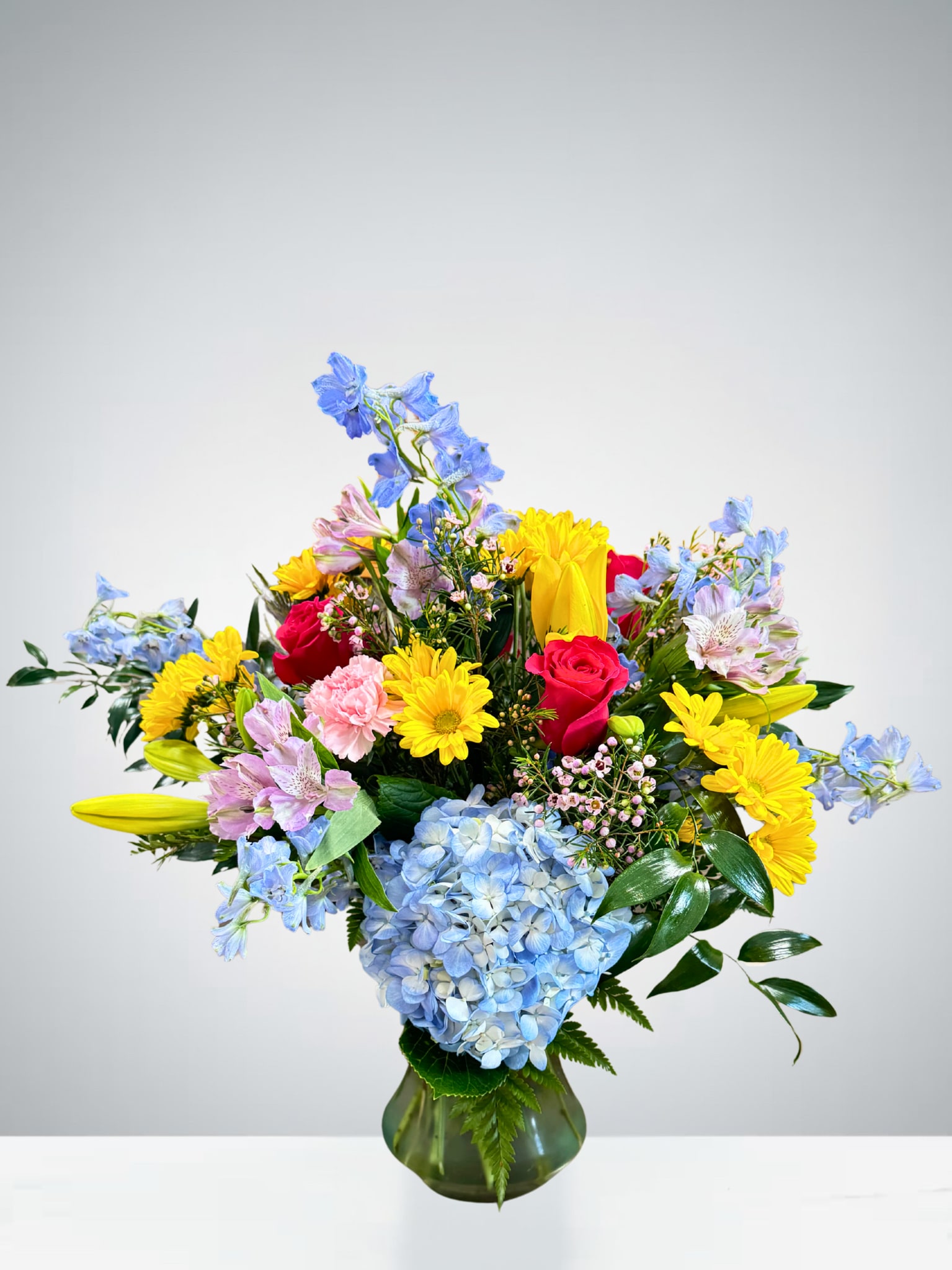 Bright &amp; Beautiful Blooms - This vibrant arrangement bursts with color and charm, featuring radiant yellow daisies, soft pink carnations, delicate blue hydrangeas, striking hot pink roses, and cheerful touches of lilies, alstroemeria, and delphinium. Arranged in a classic green vase, this design is full of life and joy, making it the perfect gift to brighten someone’s day, celebrate a special occasion, or simply bring beauty into any space.