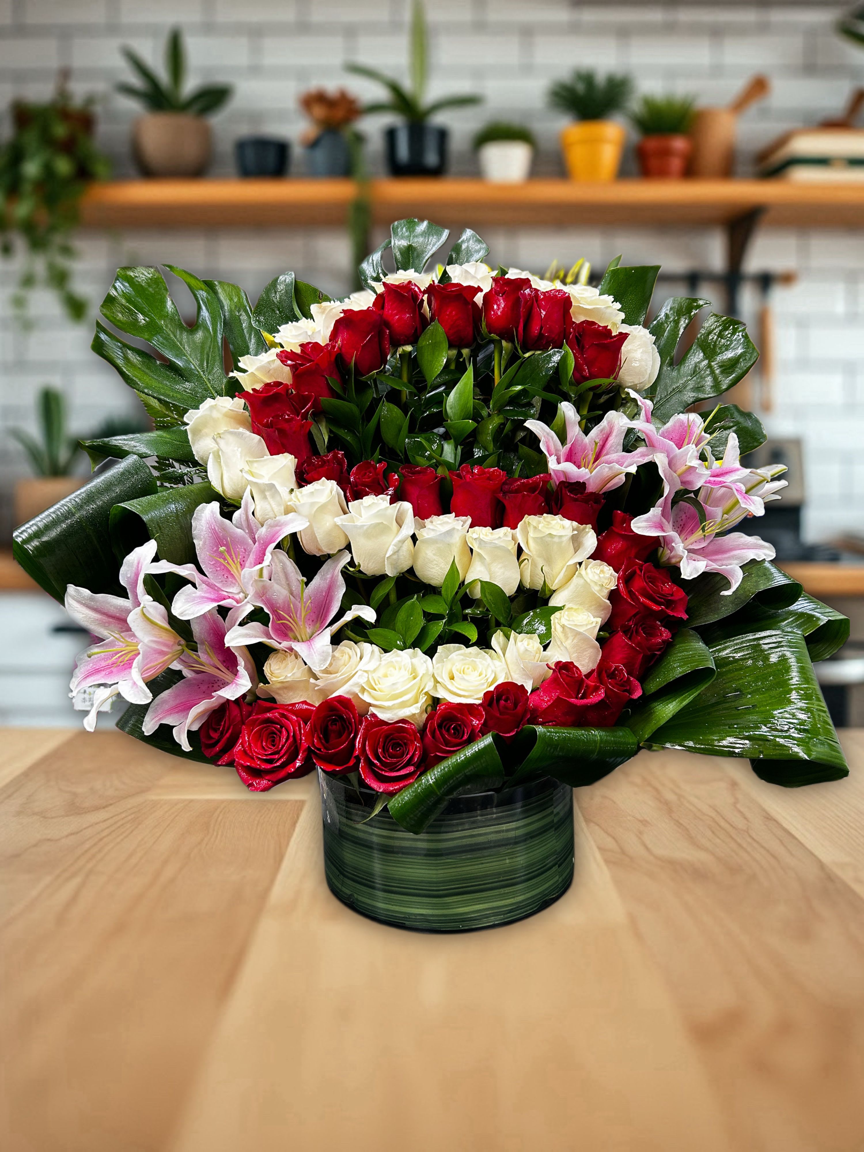 “S” letter with roses  - This is a lush and elegant floral arrangement designed in a round glass vase lined with green leaves. The centerpiece features a striking combination of fresh red and white roses, arranged in layered rings that create a bold contrast. Accents of soft pink lilies add a touch of romance and fragrance, while glossy tropical greenery frames the design, giving it fullness and balance. The overall look is both luxurious and vibrant, making it a perfect choice for celebrations, love expressions, or as a show-stopping gift.