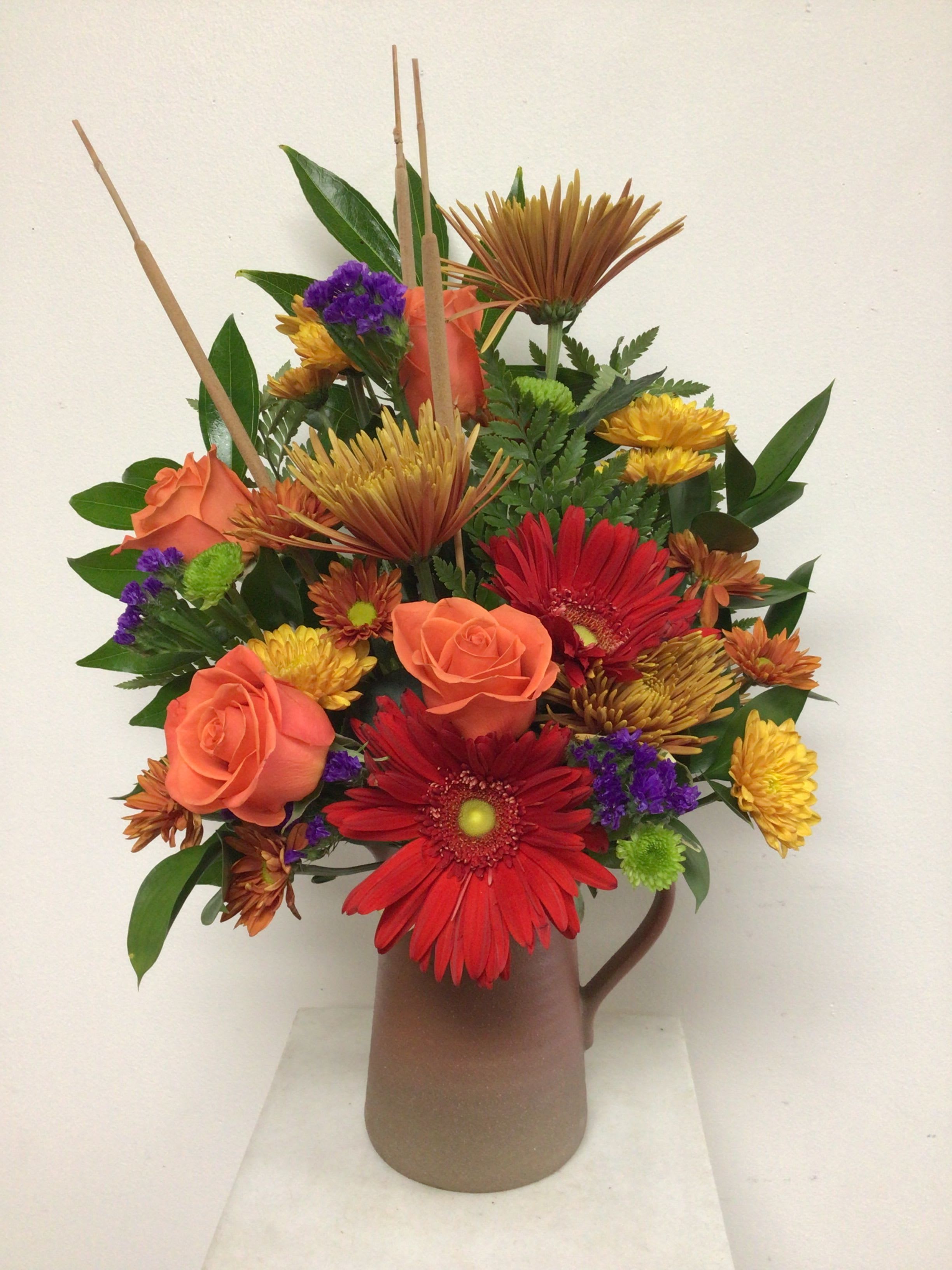 Autumn Abounds - 7 inch tall brown stoneware pitcher is arranged with orange roses, red gerbera daisies, purple statice, cattails and a colorful mixture of fall mums. This is a one sided arrangement. Approximate size is 20 inches tall by 12 inches wide.