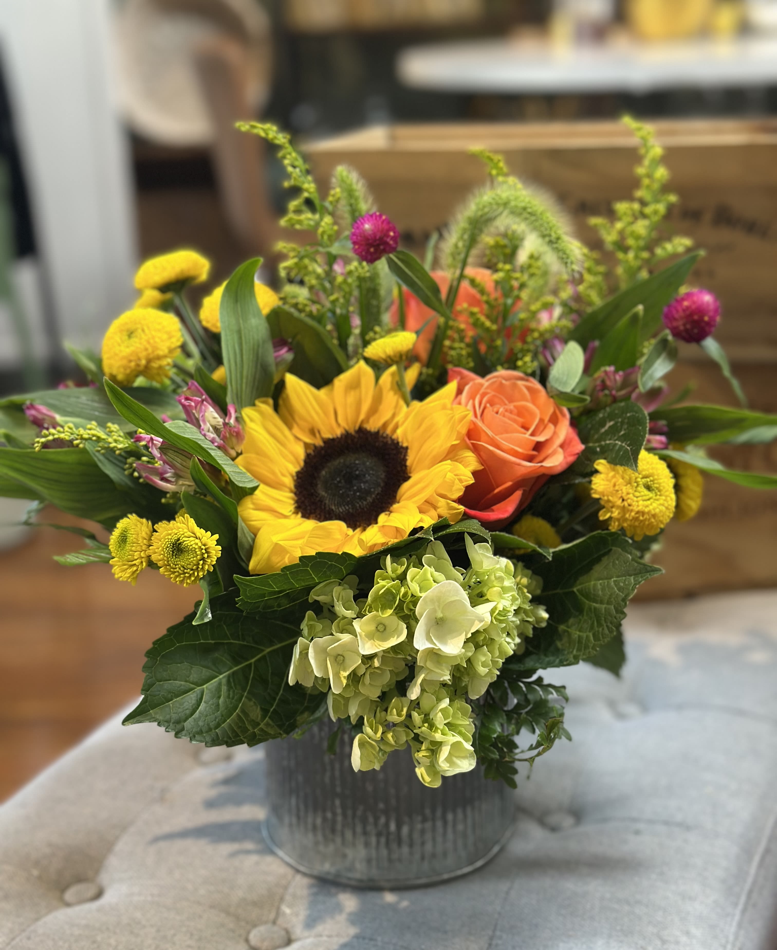 Golden Meadow - Golden Meadow is a cheerful, lush arrangement featuring bright sunflowers, and warm orange roses. Wisps of millet grass and playful gomphrena add natural texture, creating a wild, sun-kissed look in tones of yellow, orange, and pink. Inviting sunlit charm and soft elegance into any space.