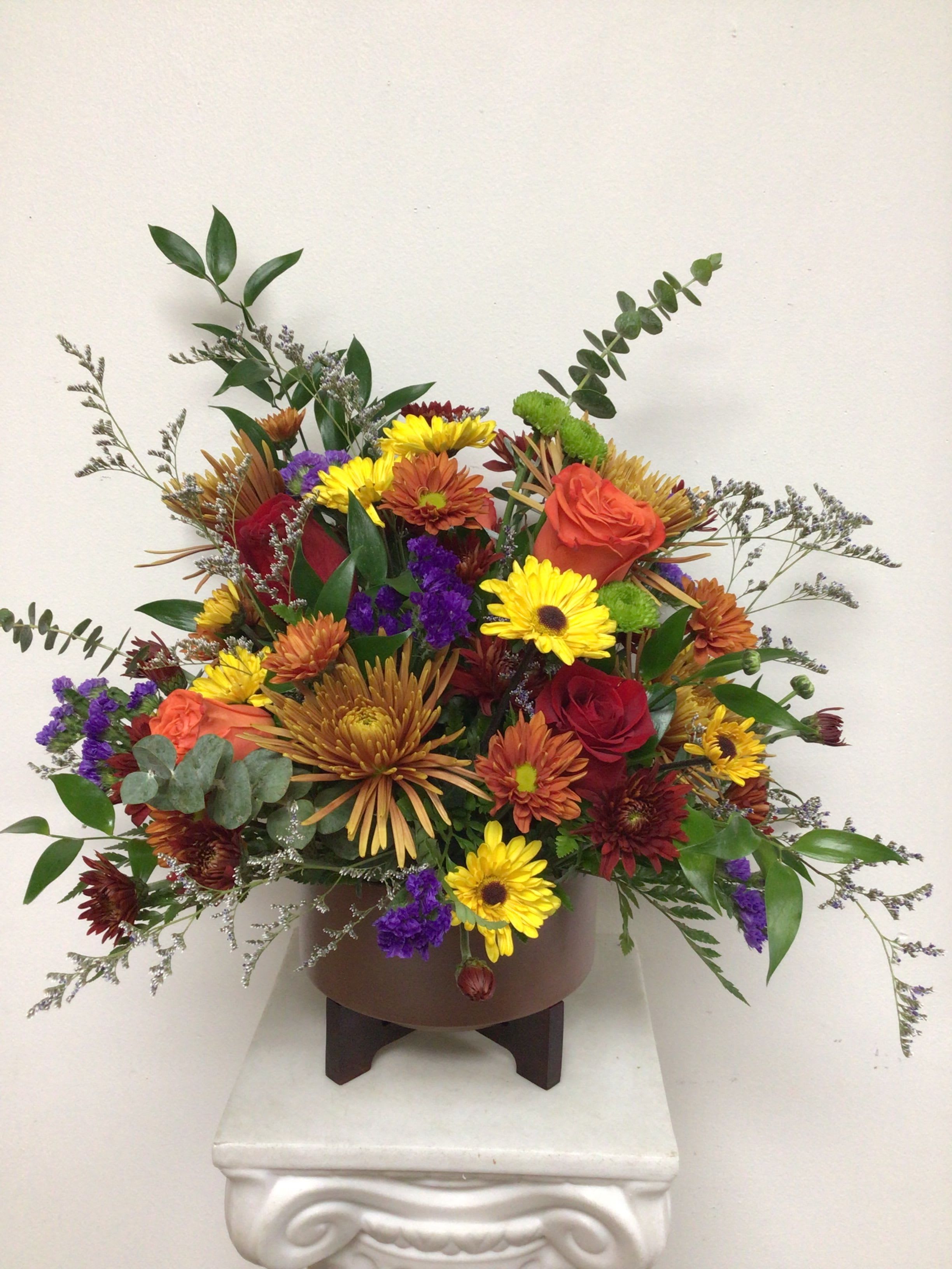 Festive Fall - Contemporary copper container is 7 inches wide, 4 inches tall on a wooden base and arranged with red, roses, orange roses, purple statice and beautiful mixed fall mums. This is an all around arrangement. Approximate size is 15 inches tall by 18 inches wide. 