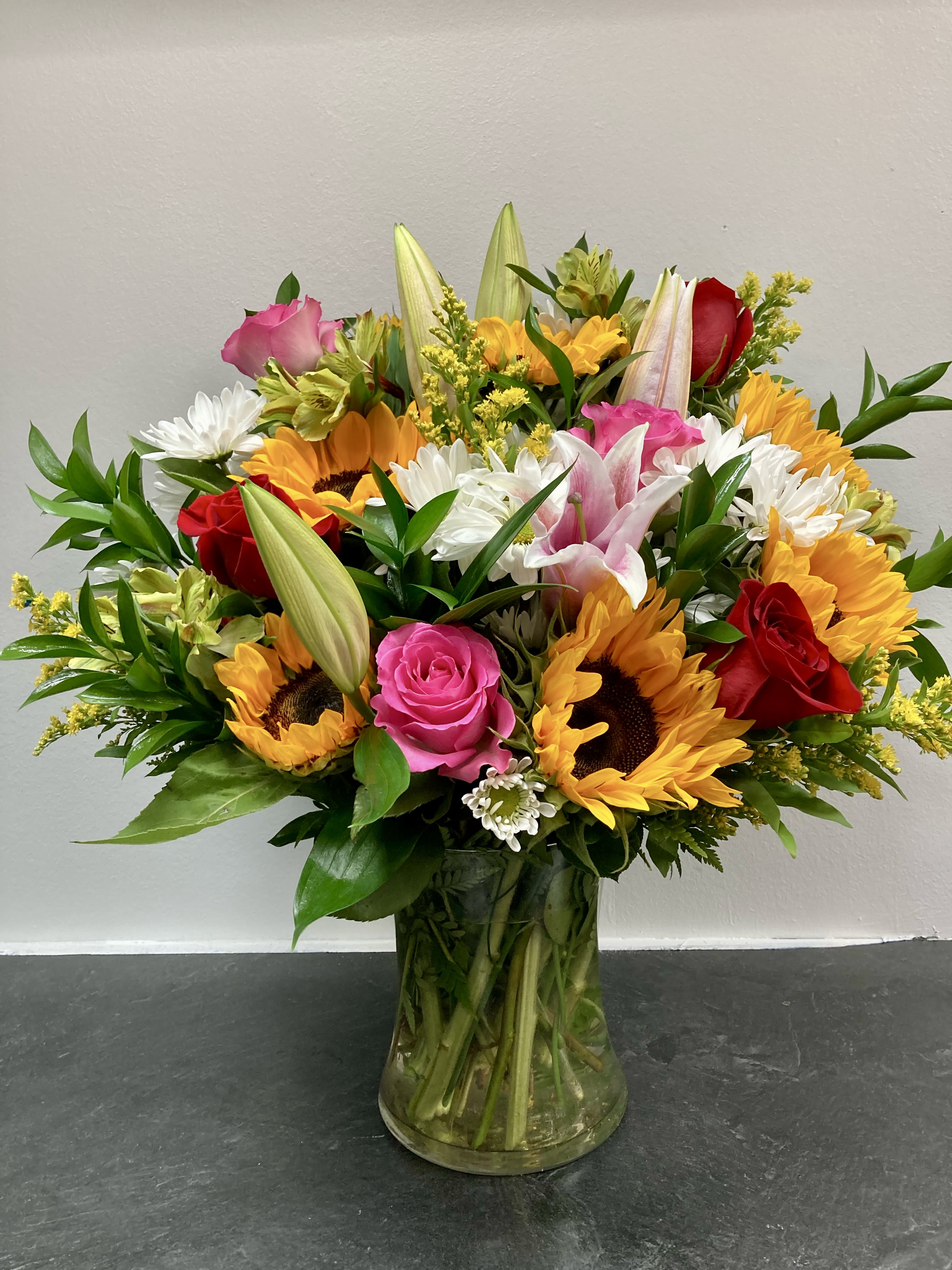 Sunshine arrangement.  - A radiant mix of cheerful sunflowers, elegant lilies, and classic roses arranged in a clear glass vase. Bright yellows, vibrant pinks, bold reds, and crisp whites come together with lush greenery to create a joyful and uplifting display. Perfect for birthdays, celebrations, or simply to brighten someone’s day, this bouquet balances warmth and sophistication with a fresh, garden-inspired charm.