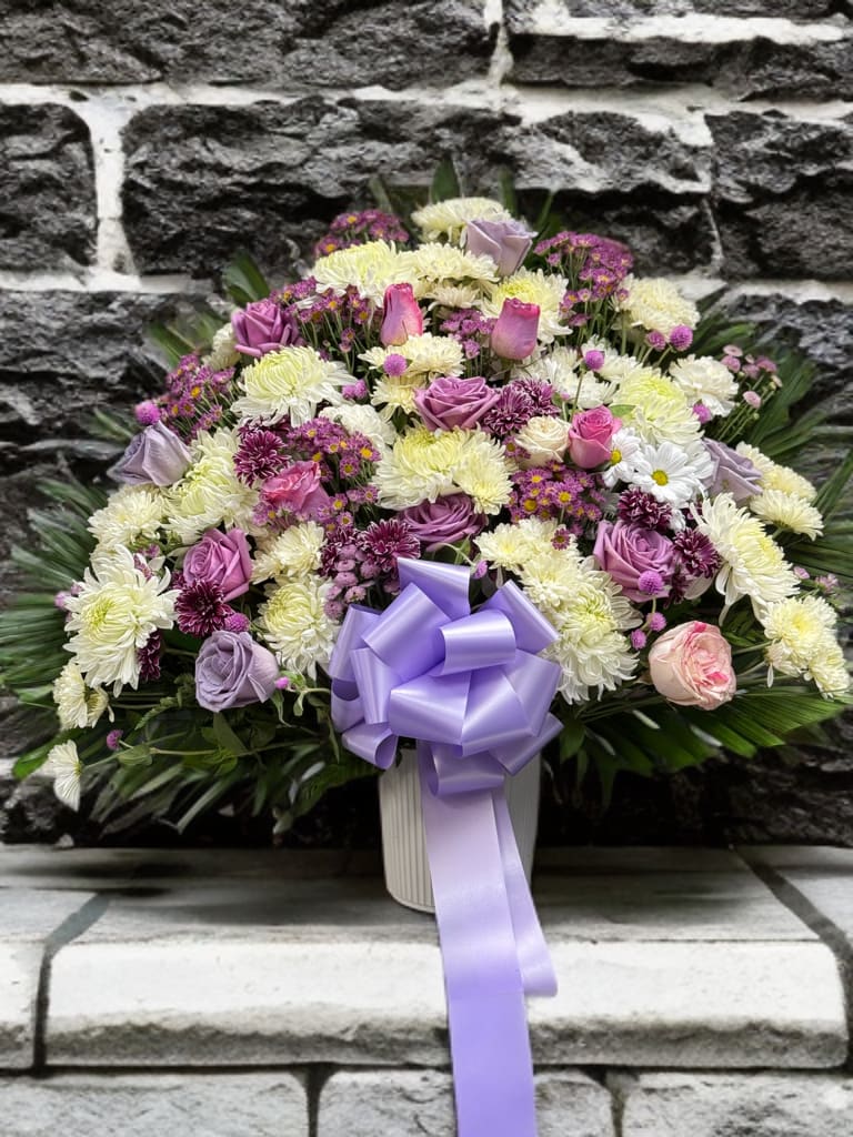 Lavender Peace Basket - This graceful funeral basket features a soothing blend of soft lavender roses, pink blooms, and delicate white chrysanthemums, accented with lush greenery and a satin lavender bow. The gentle pastel tones symbolize peace, love, and remembrance, offering comfort and serenity in times of loss. Thoughtfully designed, it serves as a heartfelt tribute that honors the memory of a loved one with beauty, dignity, and compassion.