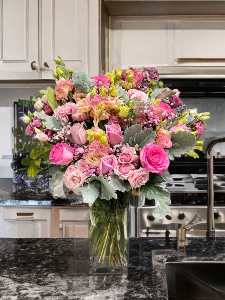 Blush Elegance Vase - A graceful arrangement of pink roses, spray roses, and soft pastel blooms accented with dusty miller and lush greenery, beautifully styled in a tall glass vase. The delicate shades of blush, hot pink, and soft peach create a romantic and uplifting display, perfect for celebrating love, birthdays, or simply brightening any space. Elegant and feminine, this bouquet radiates warmth, charm, and timeless beauty.