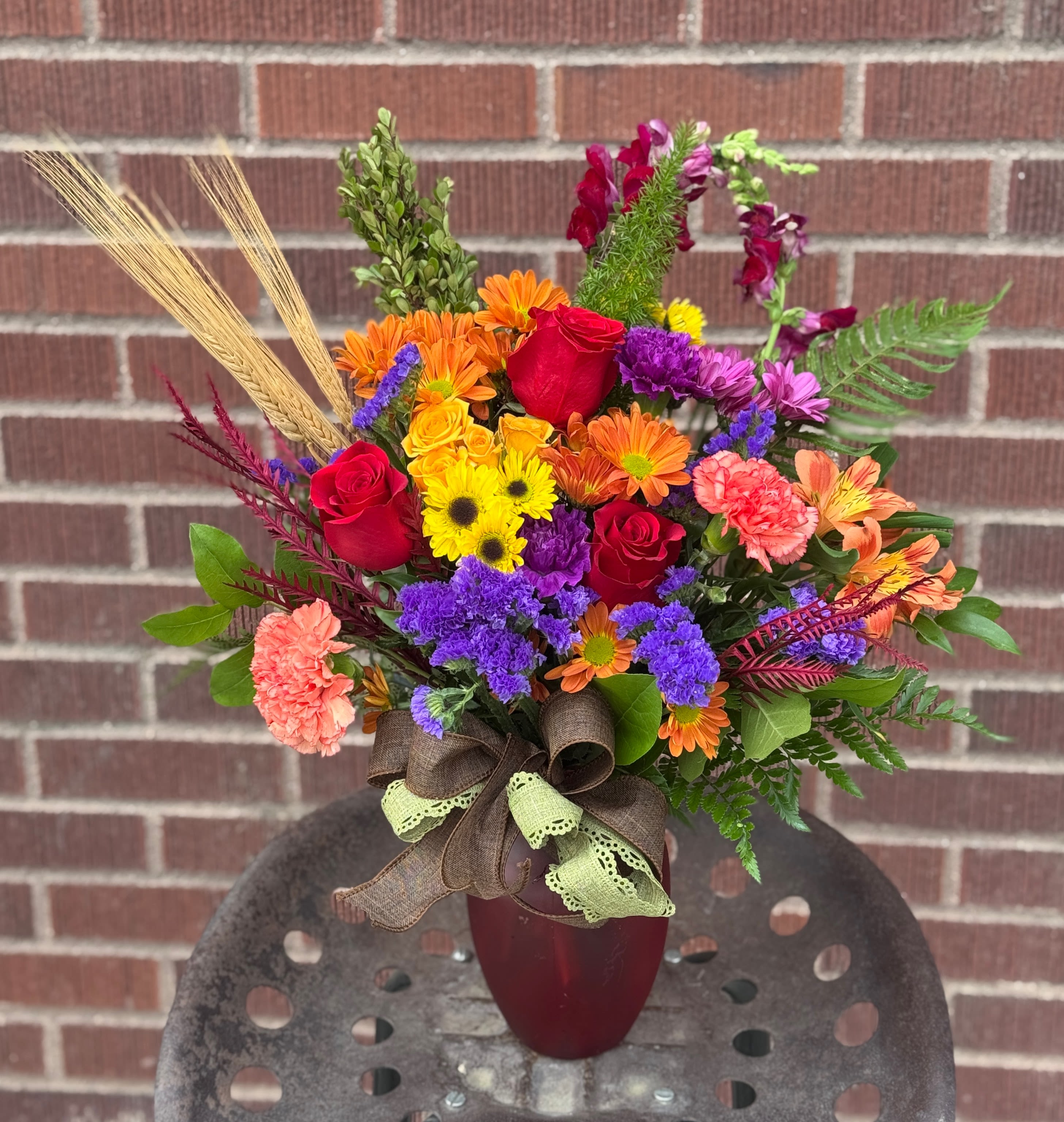 Harvest Hues Harvest Hues - Designed by one of our local gals, this piece includes wheat and burlap and the red, orange, yellow and purple tones we love when it comes to fall! Arrives in a fall colored glass vase.