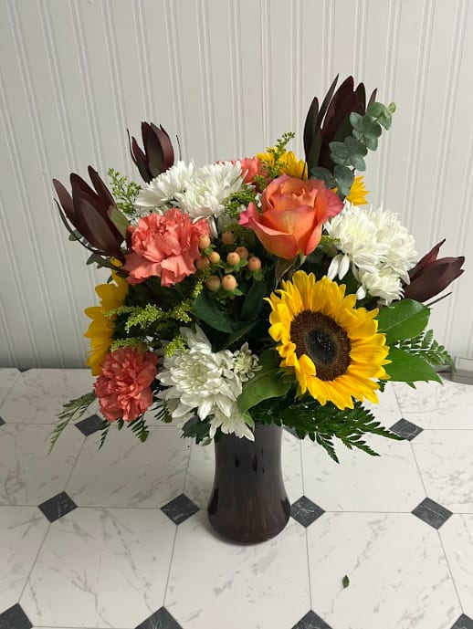 Autumn Sunset - Just like the evening sky, this arrangement blends glowing fall tones with soft, airy accents. Designed in a colored vase, it features vibrant oranges, golden yellows, and deep autumn blooms, accented by white daisies that float like clouds against a sunset backdrop. Warm, radiant, and full of seasonal charm, it’s the perfect gift or centerpiece to brighten any fall day.