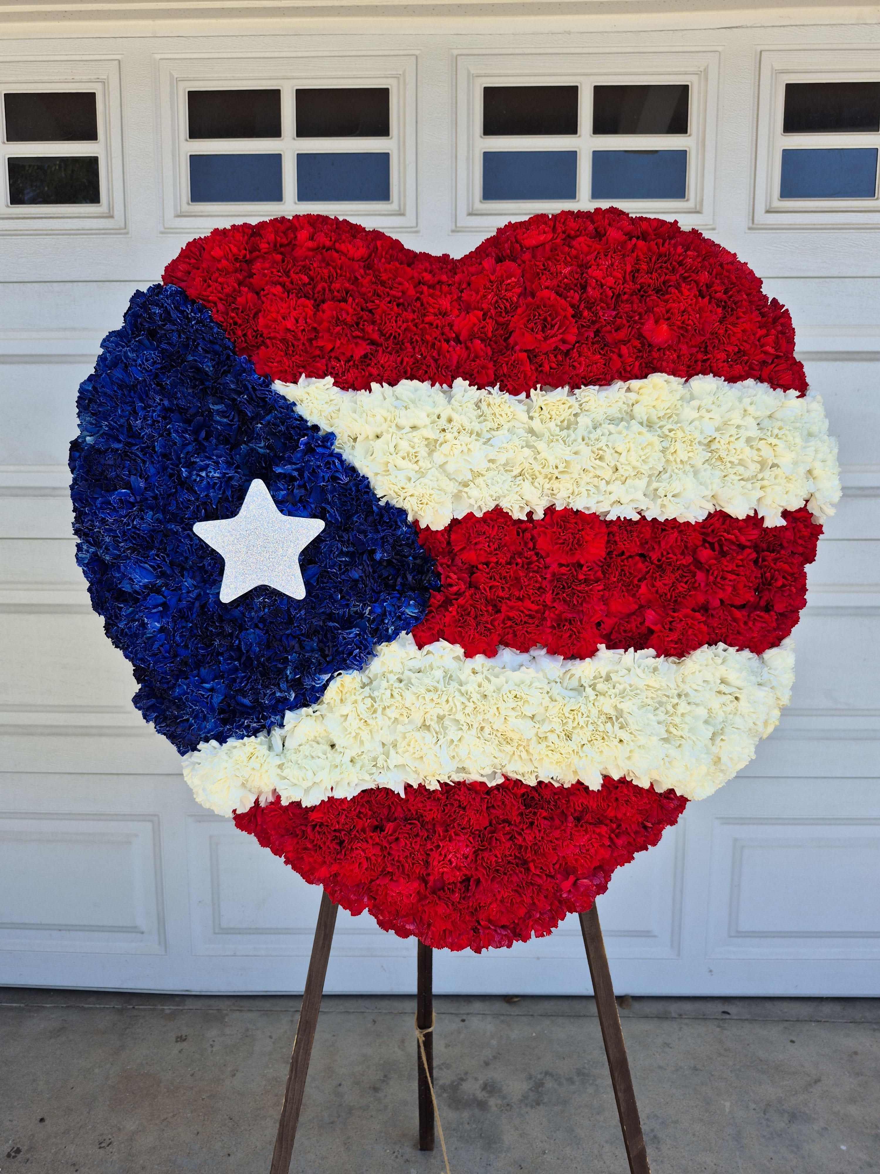 Puerto Rican Flag Standing Heart  - A perfect way to honor a heritage is by showcasing their flag at their service.  White, Red, and Blue-dyed carnations with a silver-star is brought together designed to form similarly of the Puerto Rican Flag, offering a symbol of honor and devotion at their memorial service.  The standard is without the greeneries and the deluxe is with the greenries. 