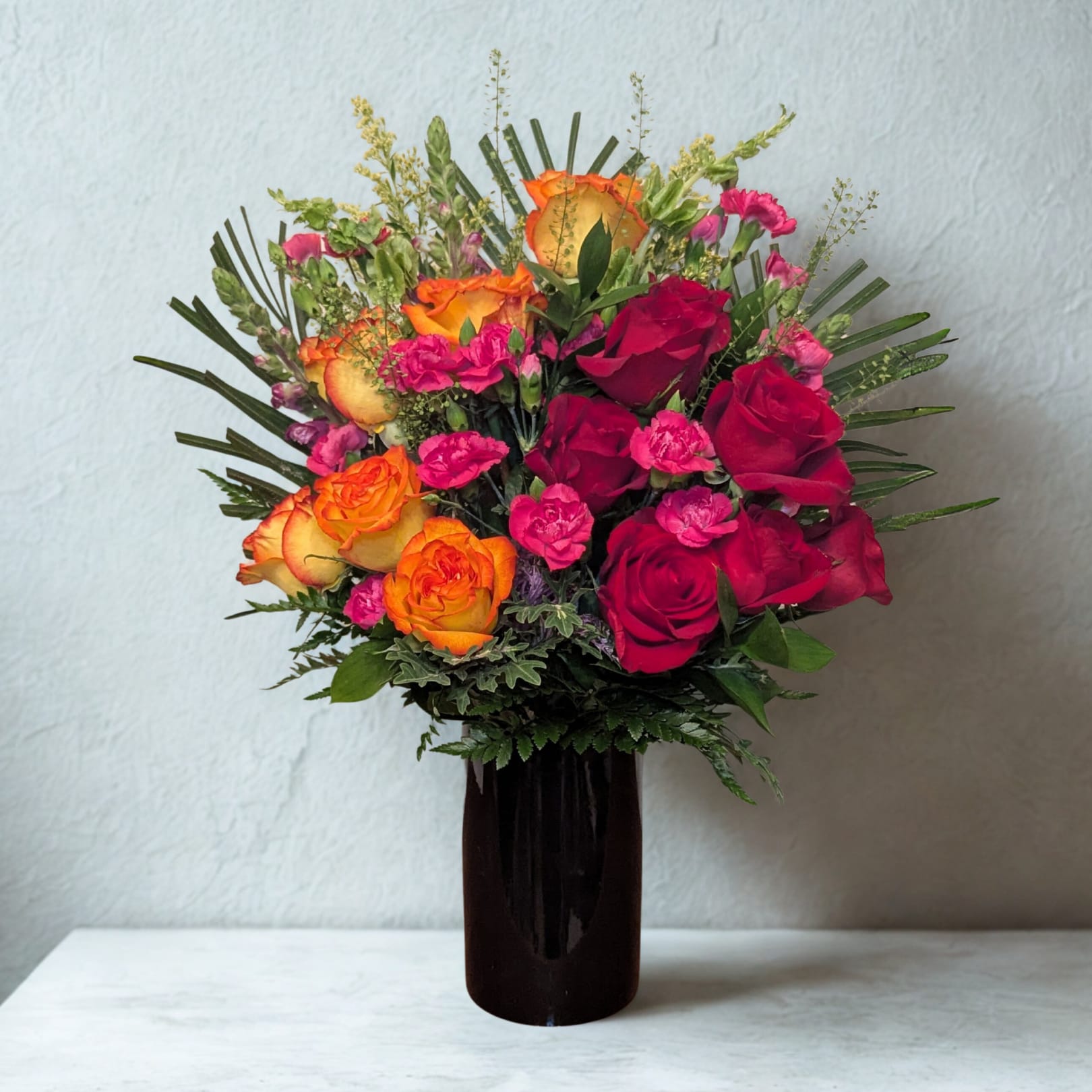 Radiant Passion Vase - A bold and vibrant expression of love and joy. This arrangement combines lush red roses with fiery bi-color orange roses, accented by playful hot-pink mini carnations, airy snapdragons, and delicate greenery. Designed in a sleek black vase, it’s perfect for birthdays, anniversaries, or simply to brighten someone’s day with elegance and warmth.