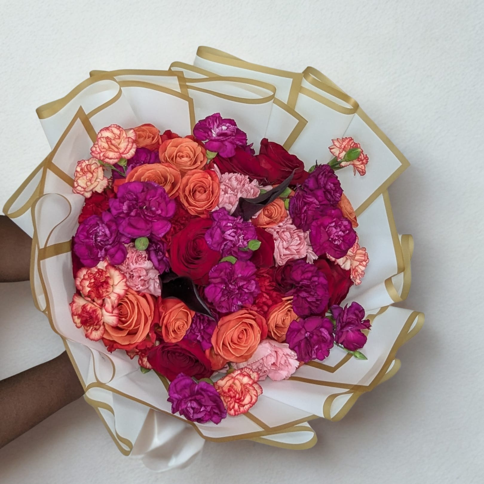 Crimson Sunset Bouquet - A bold hand-tied bouquet that radiates elegance and passion. Featuring deep red roses, vivid orange spray roses, lush hot-pink and magenta carnations, all wrapped in premium white and gold-trimmed paper with a satin ribbon finish. Perfect for birthdays, celebrations, or a romantic surprise this bouquet makes a statement before it’s even placed in a vase.
