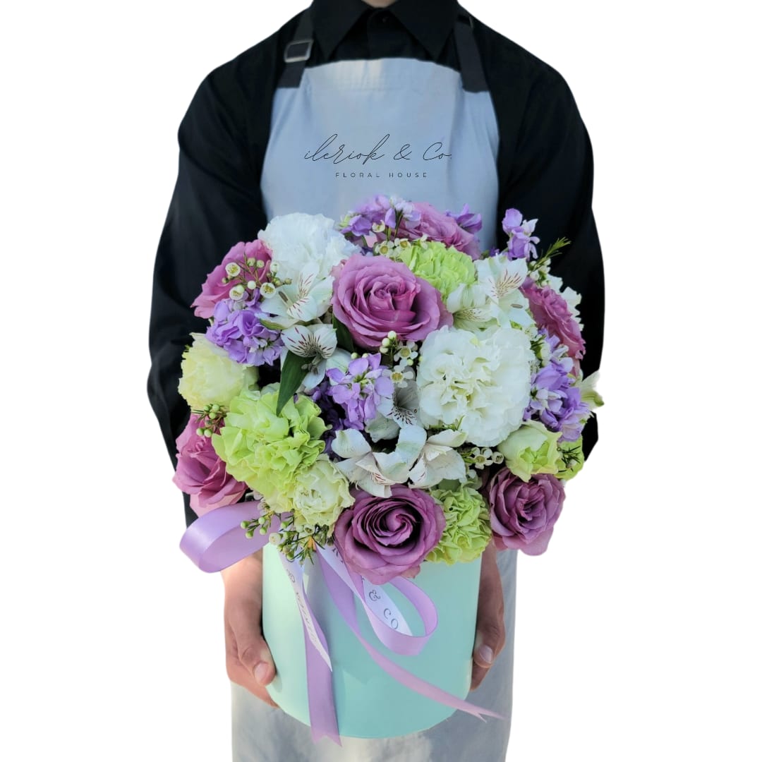 The Mimi Floral Box - Unique just like them.  A combination of purple roses, green carnations, lisianthus, stock and wax flower. In a teal floral box.   Deluxe Box Pictured