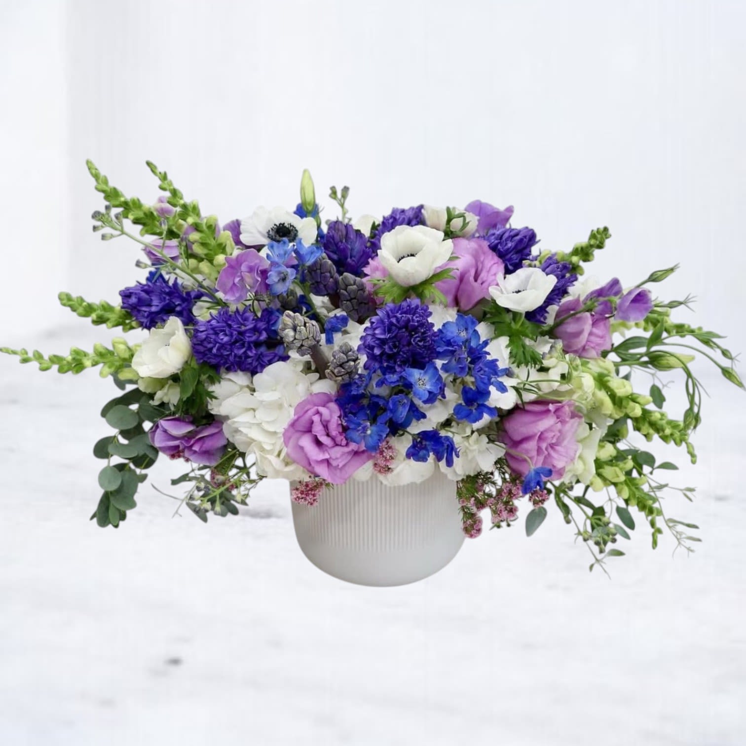 Ultraviolet Rush Ultraviolet Rush - Ultraviolet Rush is a striking floral statement designed for those who love vibrant, unexpected beauty. Bursting with electric shades of indigo, violet, and crisp white, this arrangement features lisianthus, delphinium, anemones, hyacinths, snapdragons, and seasonal textures, artfully arranged in a sleek white ceramic vase.