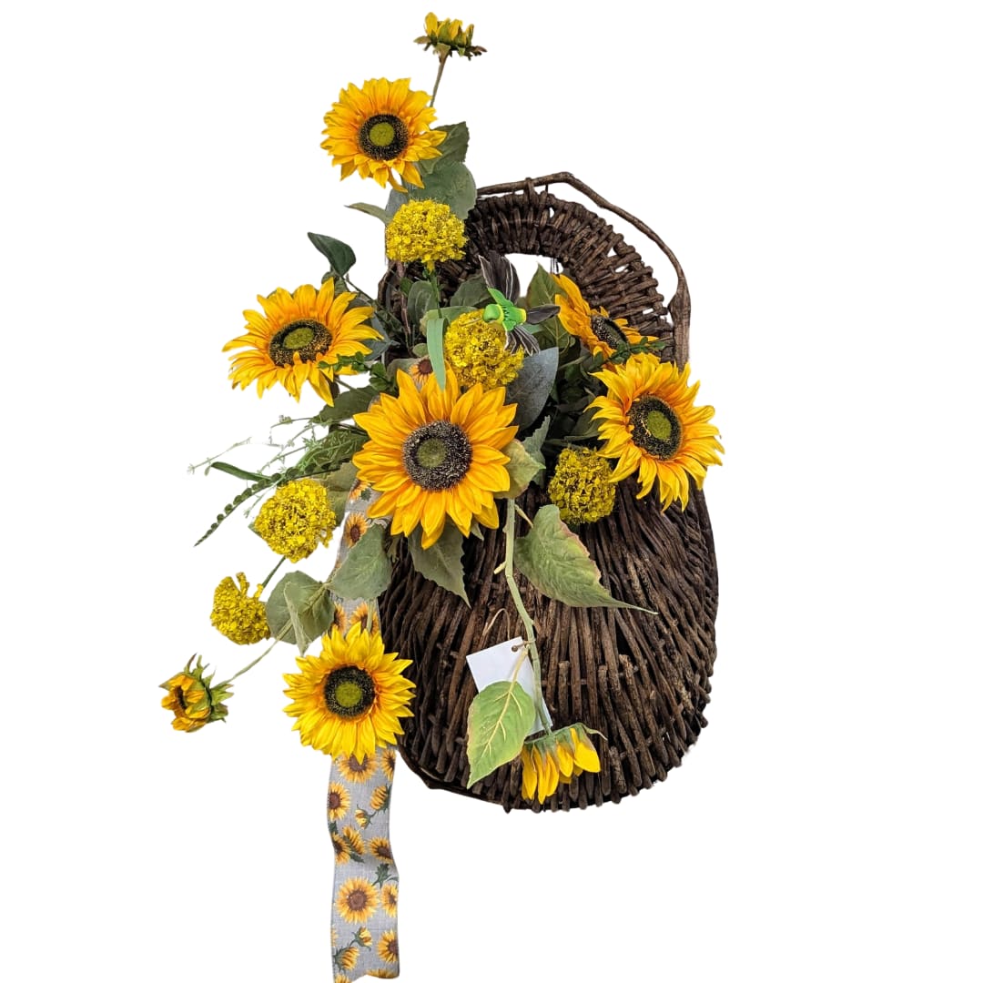 Sunlit Serenity Wall Basket  - Elevate your space with the captivating charm of the Sunlit Serenity Wall Basket. This exquisite arrangement features vibrant silk sunflower stems artfully nestled within a dark wicker wall basket, offering a delightful contrast that enhances its natural beauty. The ensemble is elegantly finished with a sunflower-patterned linen ribbon accent, adding a touch of rustic elegance to your decor.   Perfect for brightening up living rooms, hallways, or entryways, this arrangement is a timeless piece that brings a warm, sunlit ambiance to any setting.