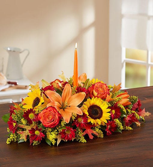 Traditional Fall Centerpiece - All-around centerpiece arrangement with autumn-colored roses, sunflowers, red daisy poms, orange Asiatic lilies and solidago; accented with assorted greenery and dried oak leaves. Complete with orange taper candles. Arrangements are hand-designed so colors and varieties may be substituted with similar items based on local availability. 