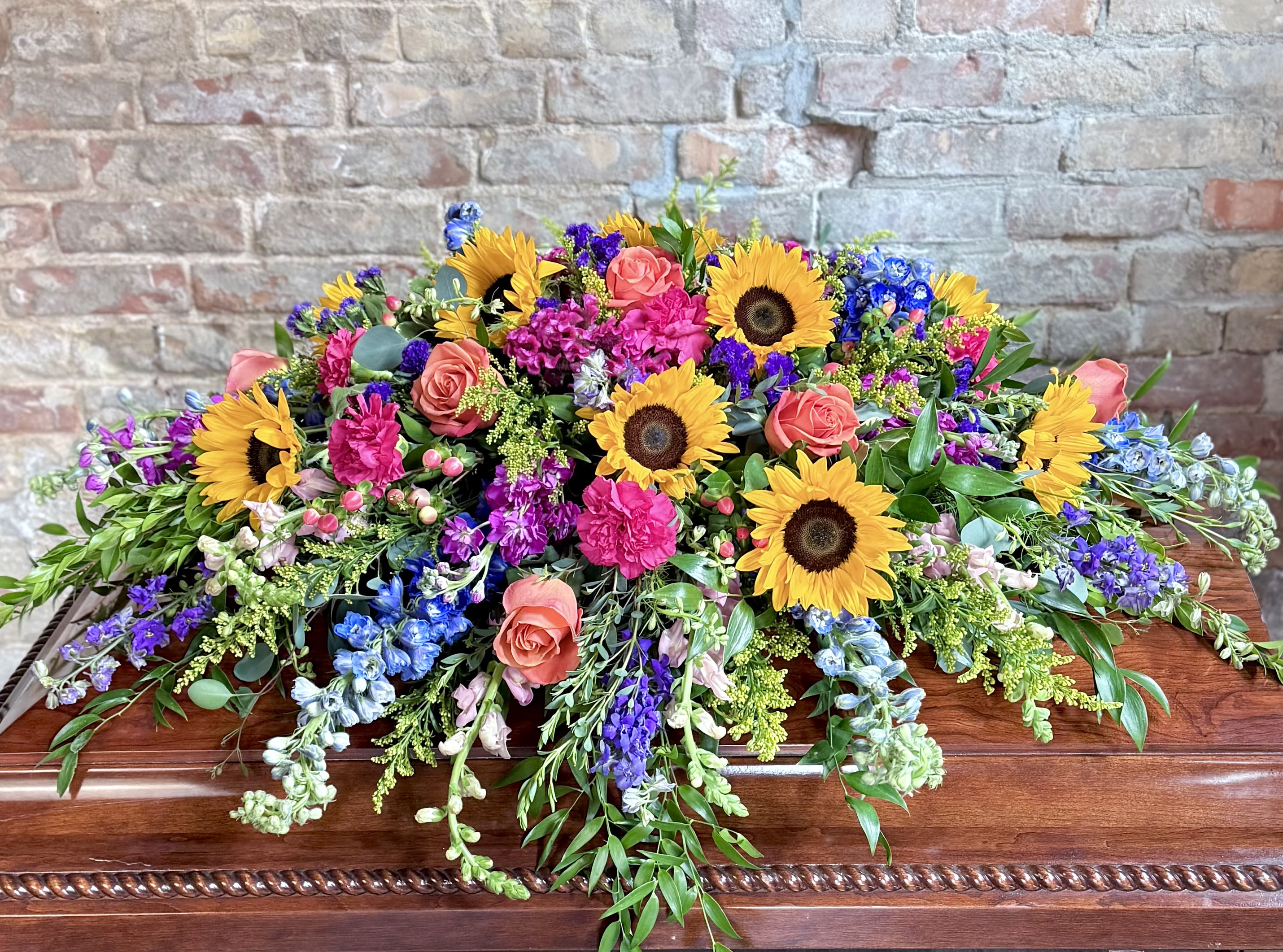Summer Sunflower Casket Spray Summer Sunflower Casket Spray - A bright and bold tribute filled with sunflowers, delphinium, larkspur, roses, berries, stock and carnations to name a few! A perfect remembrance for someone who loved bright colors and summer gardens!
