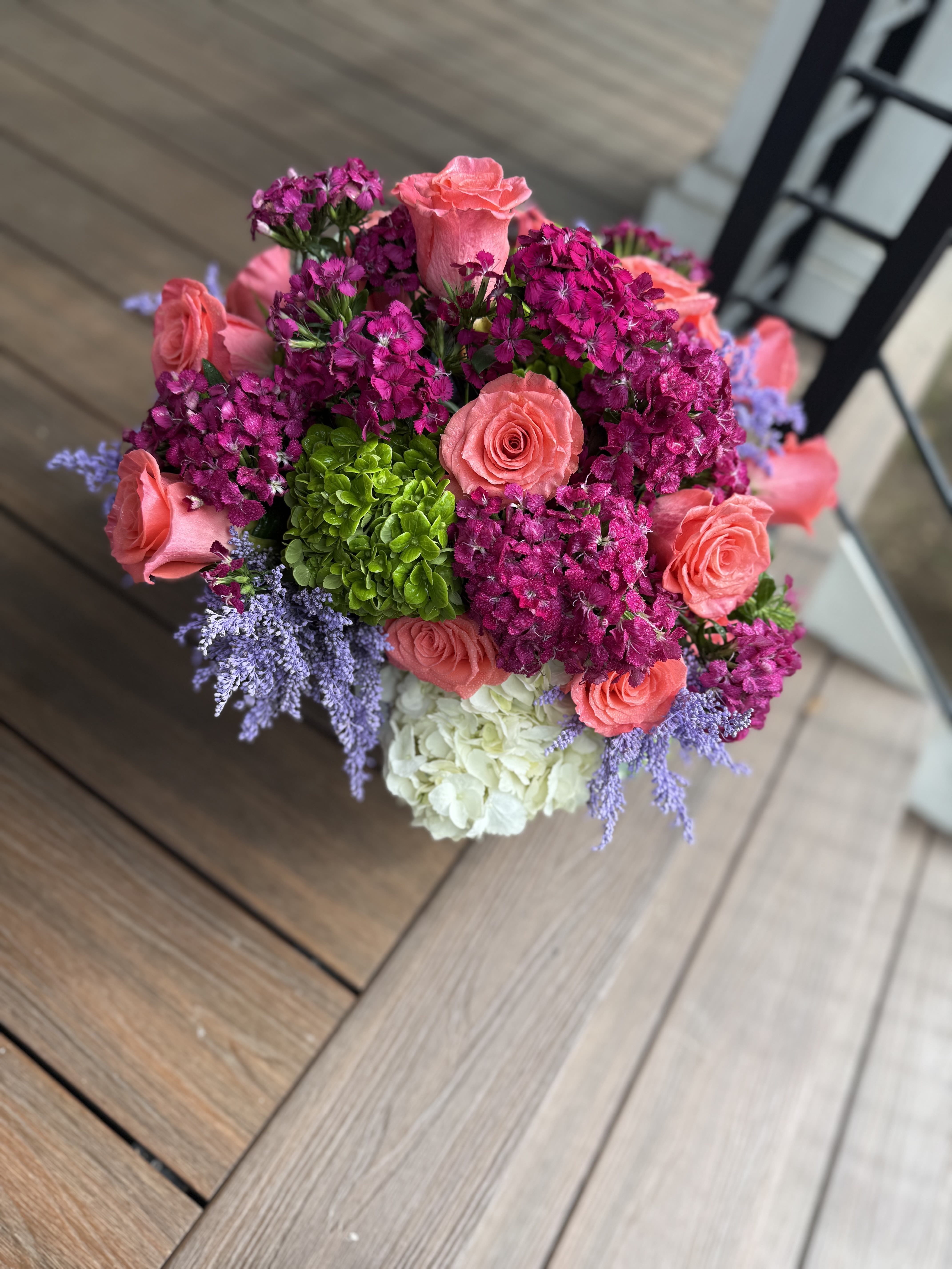 Vibrant Things - A lively and eye-catching arrangement designed to bring joy and energy to any space. This bouquet features lush roses in shades of coral, paired with antique and traditional hydrangeas for timeless elegance. Rich filler blooms in vibrant purples and greens add layers of texture and depth, all thoughtfully arranged in a sleek cylinder vase. A perfect blend of modern charm and classic beauty, this piece truly lives up to its name.