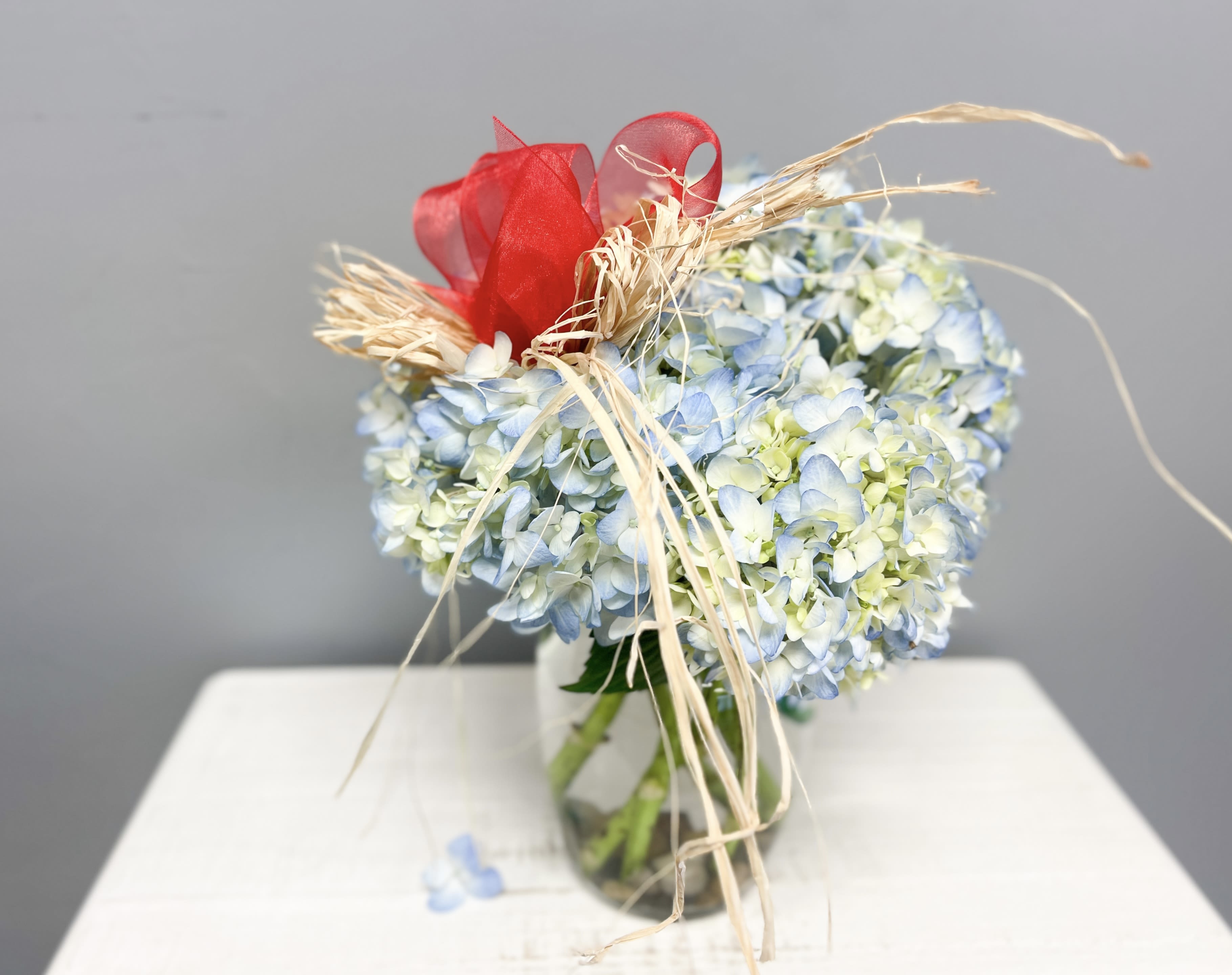 Turning Point - Turning Point” pays tribute to the life and legacy of Charlie Kirk — a figure who challenged young people to stand courageously for their beliefs and reimagine the direction of their futures. This arrangement brings together serene blue hydrangeas paired with a vibrant red ribbon and rustic raffia accents.  It’s more than a floral piece: it embodies resolve, patriotism, conviction, and the moment when one decides that change is essential. Let Turning Point be a symbol of remembrance, inspiration, and the hope that bold ideas and faith in one’s values can light the path forward.