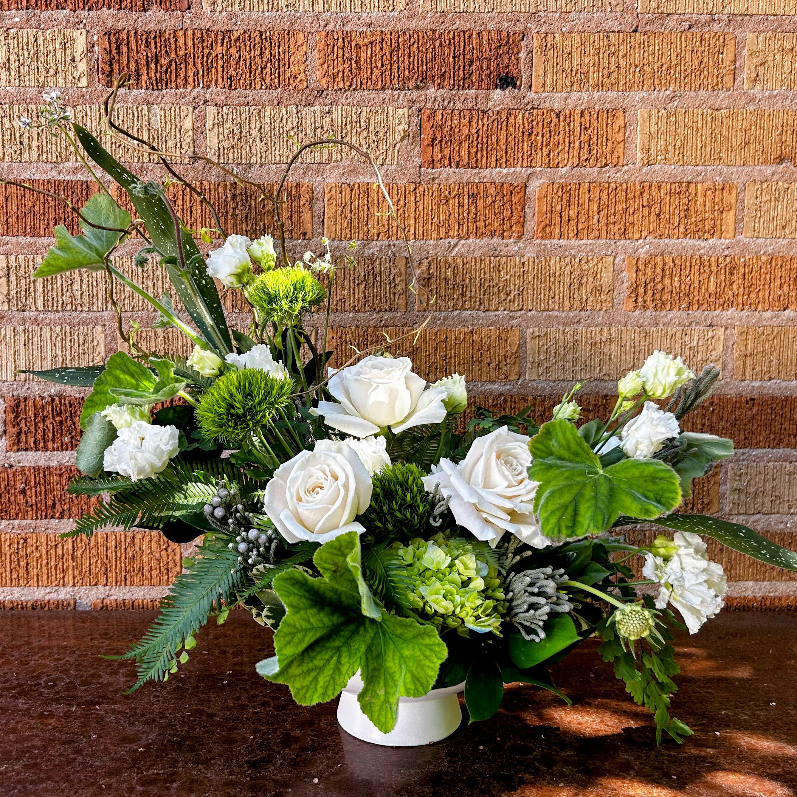 Esmerelda - Fresh, modern, airy garden style arrangement in immaculate monochrome, with a sultry half-moon shape, featuring: Playa Blaca rose, mini green hydrangea, green ball dianthus, white lisianthus, geranium, boronia, and curly willow and luxurious tropical greens.
